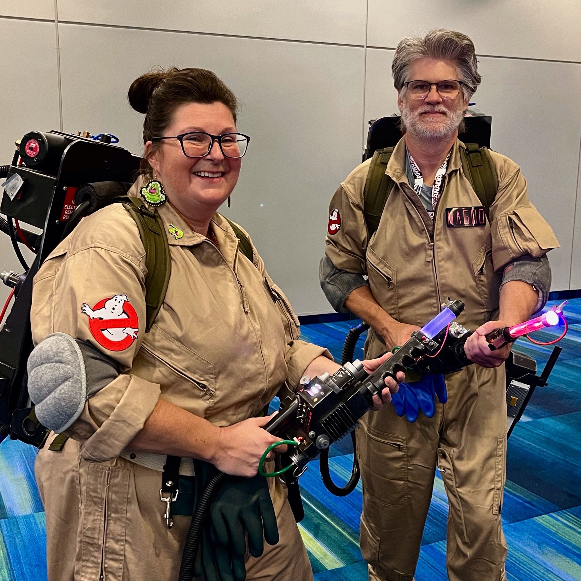 A smiling middle-aged white woman and silver-bearded white man dressed in Ghostbusters jumpsuits, particle accelerators charged!