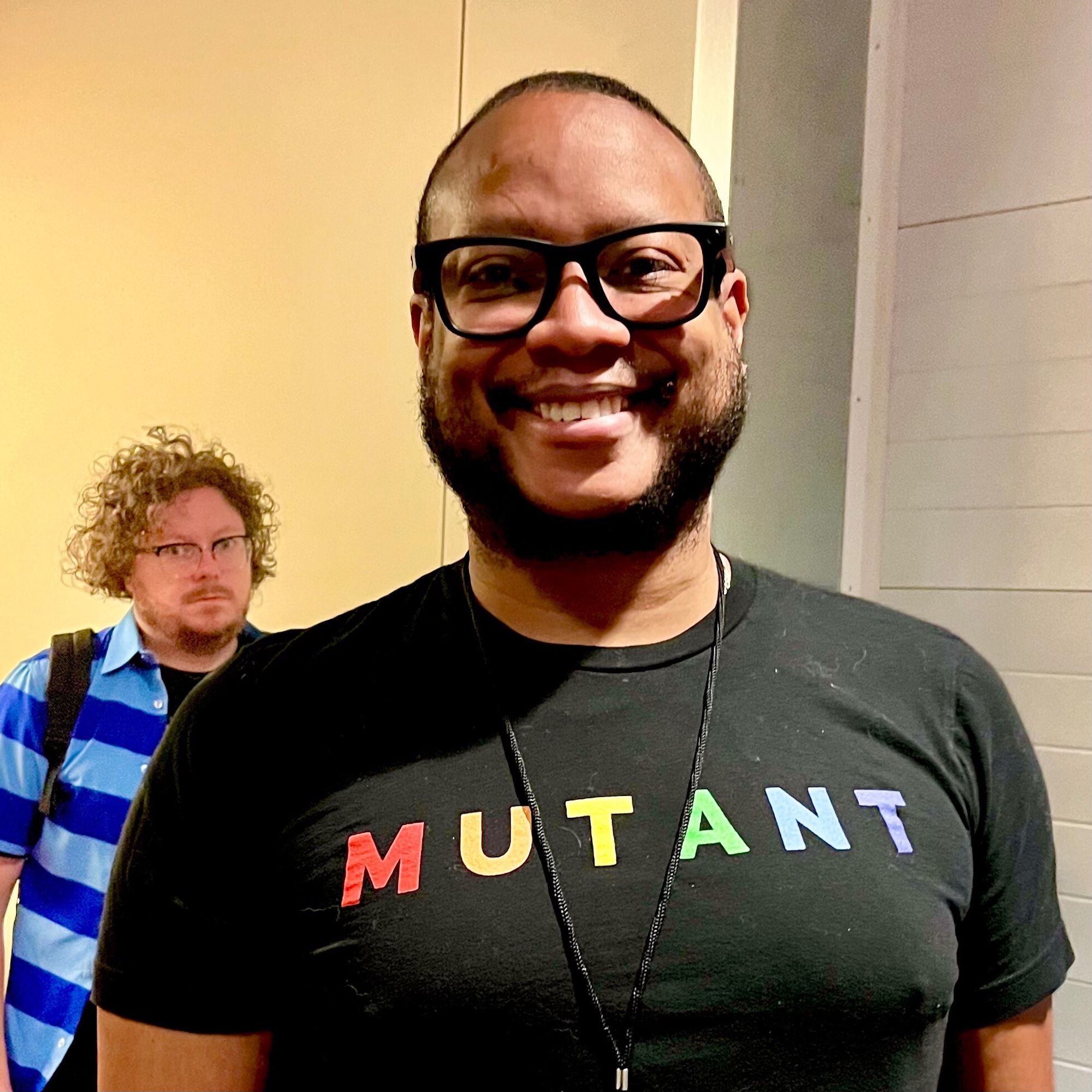 A smiling Black man in a black T-shirt with MUTANT in rainbow lettering