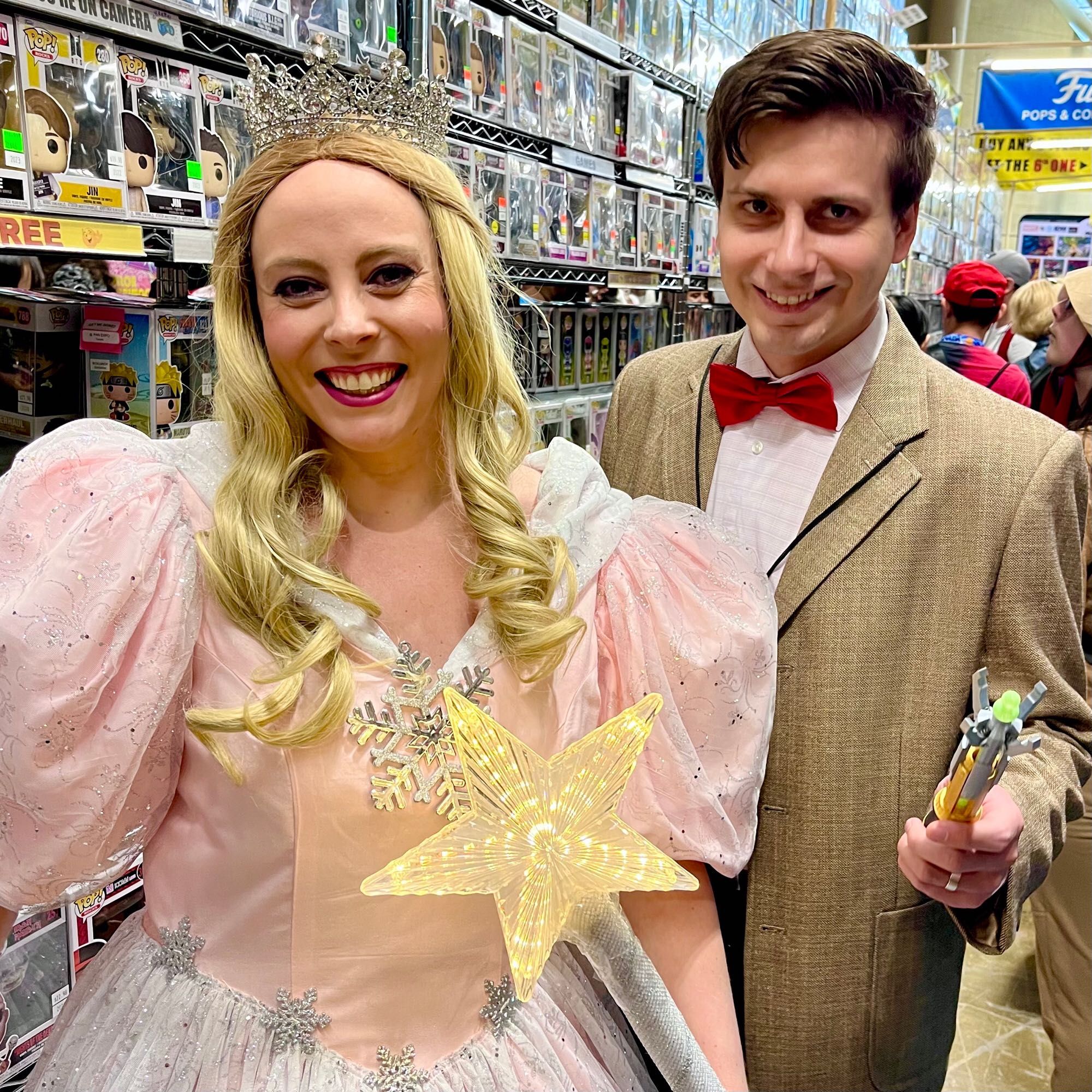 A happy young white couple dressed as Glinda the good witch with her light-up magic wand and the Eleventh Doctor with his sonic screwdriver