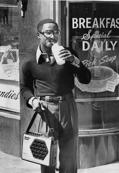 A young Black man in pleated pants, a sweater, a large-collared shirt and glasses drinks from a straw while holding a radio on a strap with his right hand. He is outside a Horn & Hardart location, advertising a daily breakfast special and rich soup.