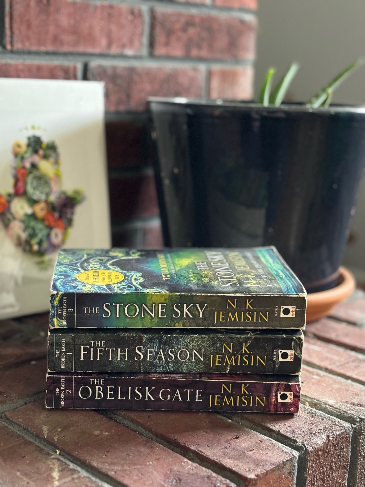 Image of NK Jemison’s Broken Earth Trilogy (Fifth Season, Obelisk Gate, Stome Sky), stacked on a brick fireplace with a dark blue pot and flower print in the background.