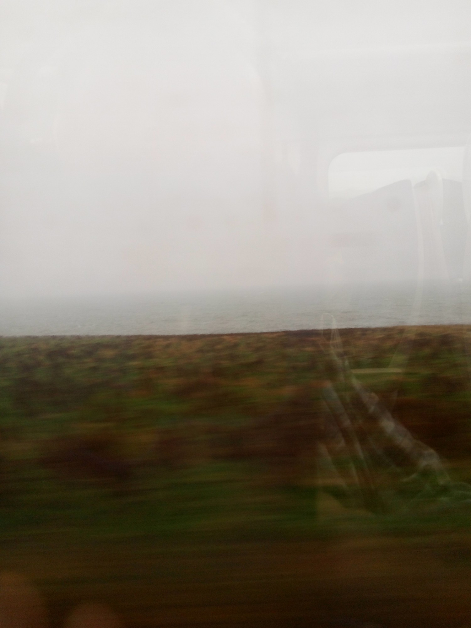 View from train window showing a field and dark grey heavy skies above a misty and grey sea