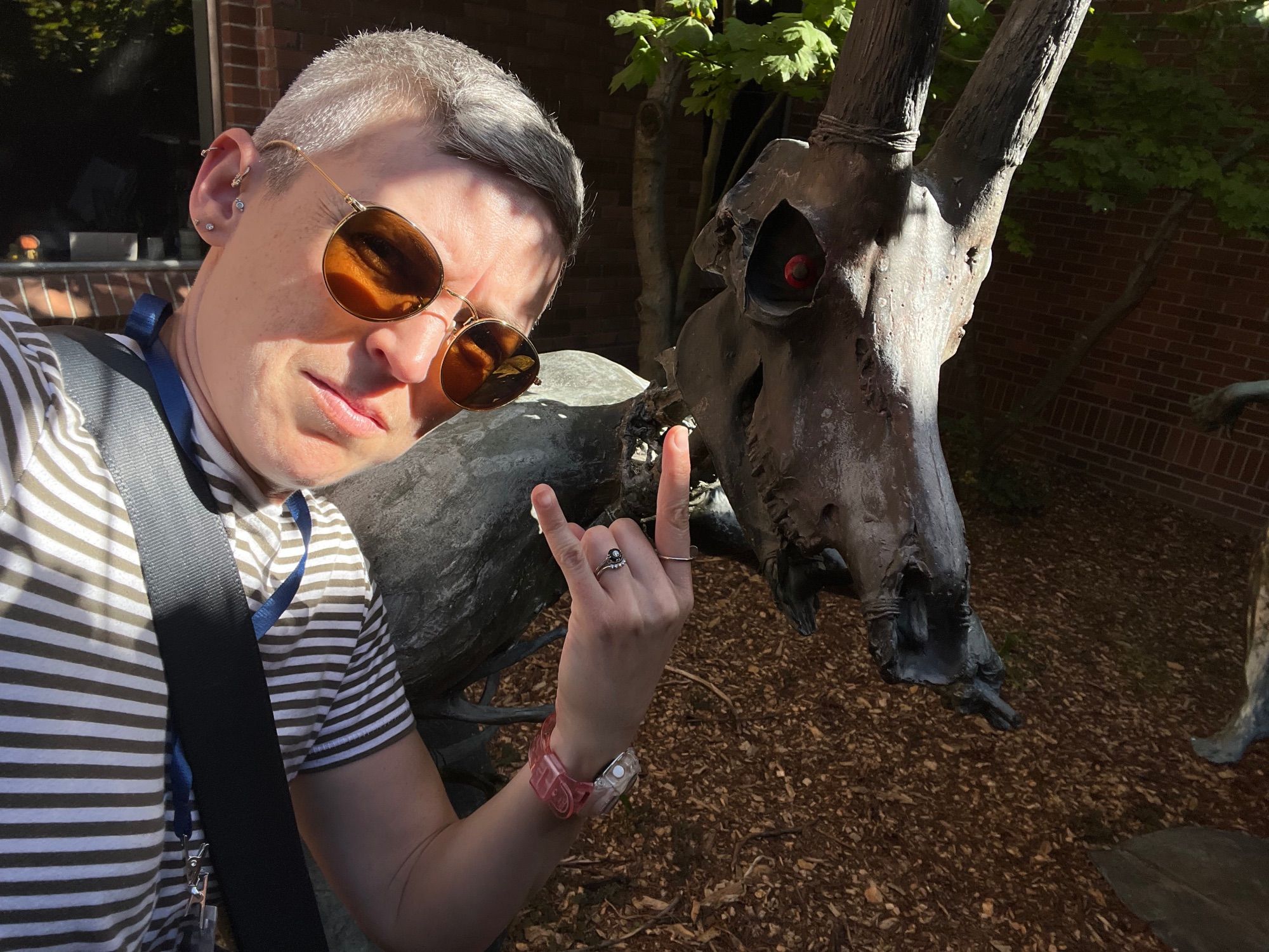 White woman in sunglasses throws horns next to a metal statue of an animal skull with a baleful red eye