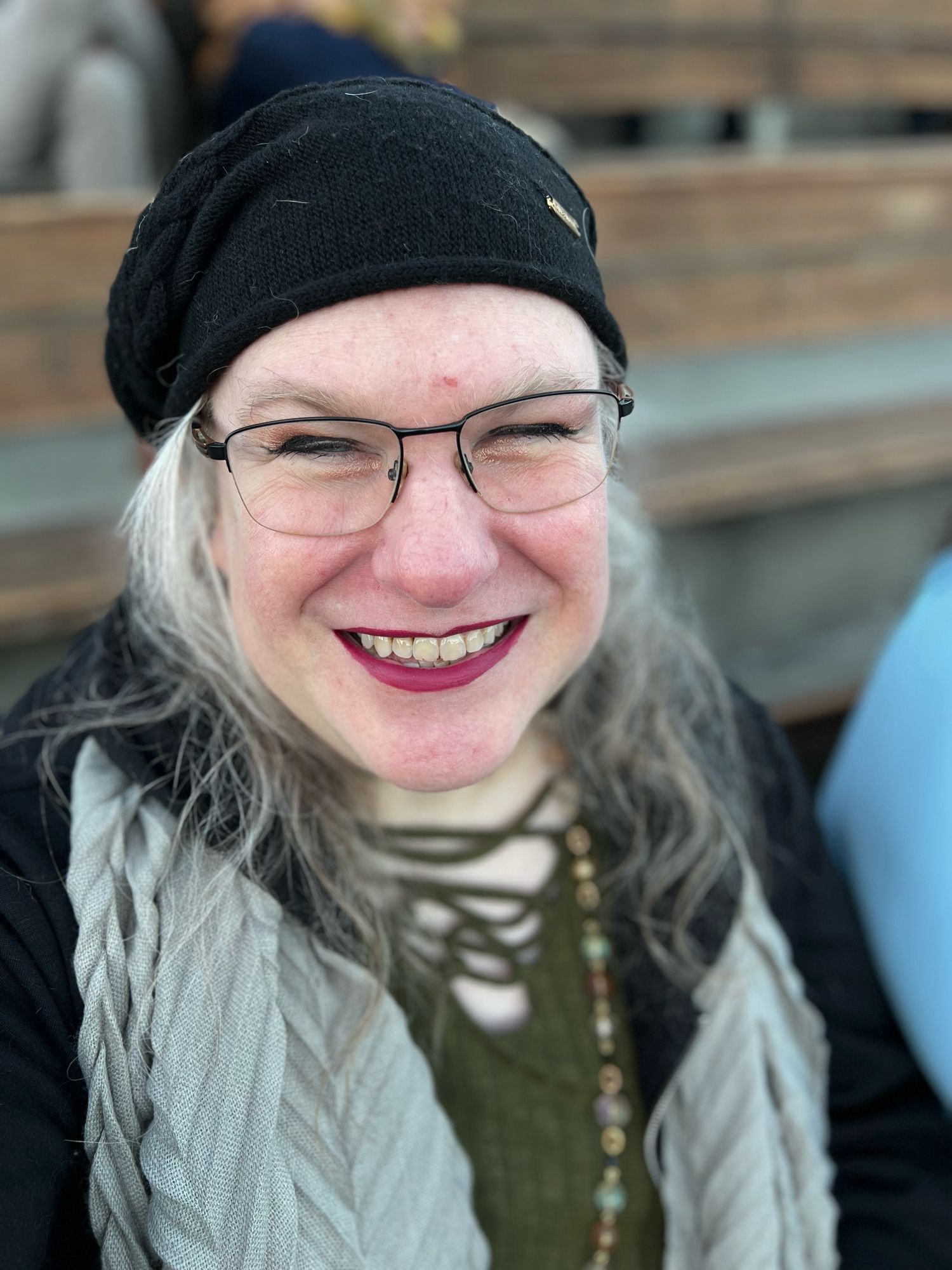 trixie sitting in an amphitheater, wearing a black hat, black hoodie, green lace-up blouse, and pale green scarf