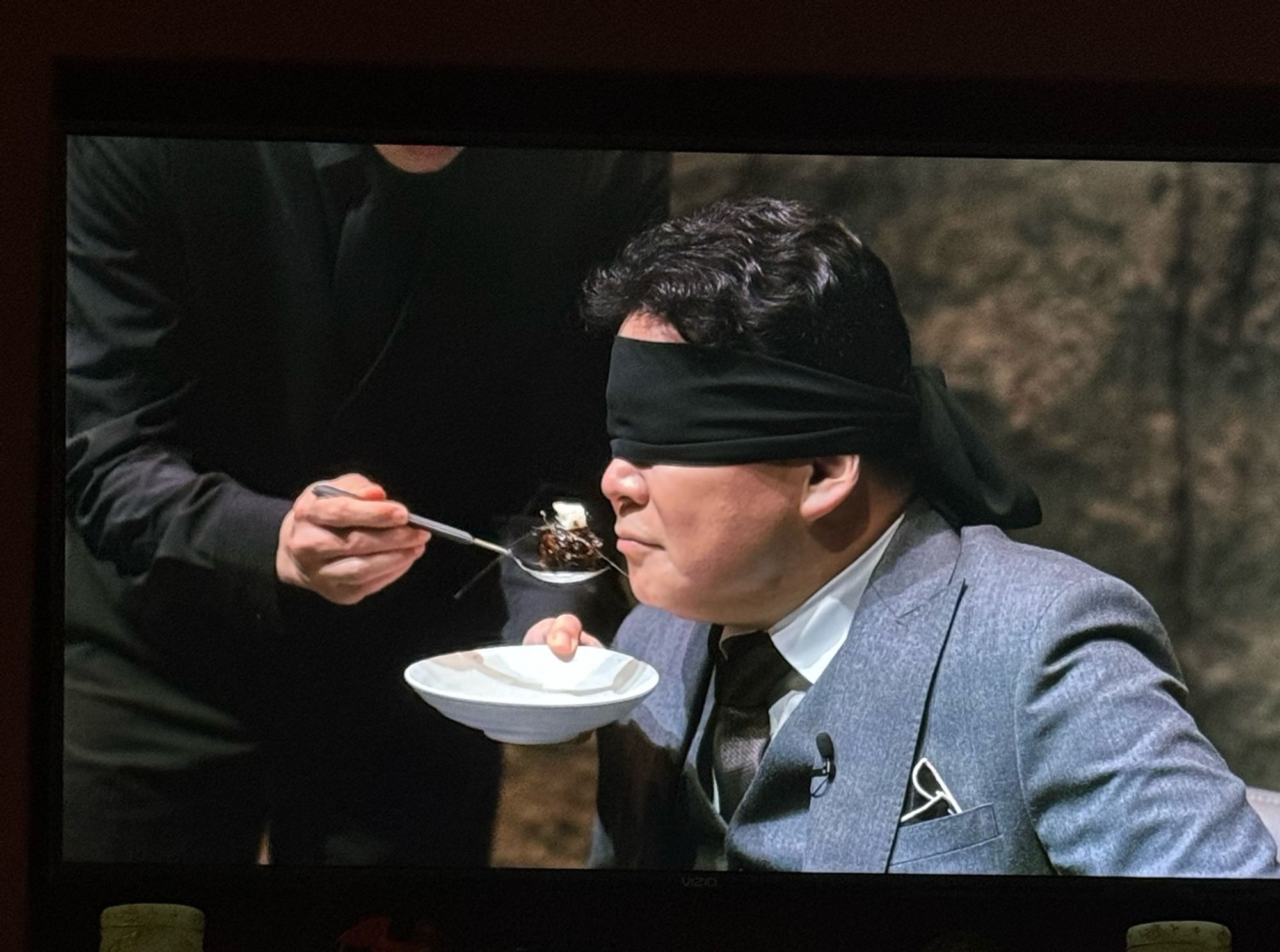 a blindfolded man in a suit being fed a spoonful of food by a waiter