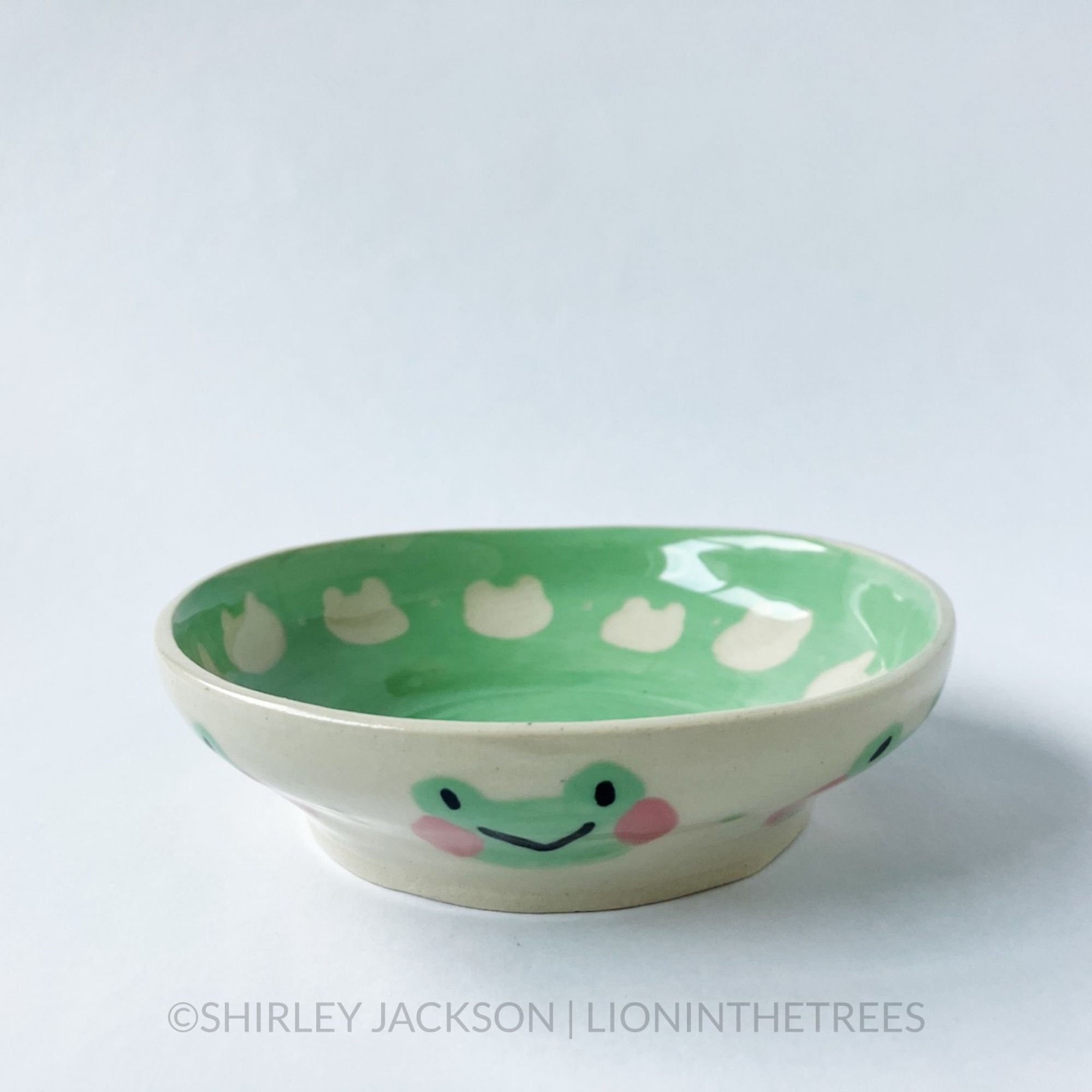 A little bowl that has a cute green frog motif on the outside done with green underglaze and a blushing pink underglaze for it's cheeks.
The interior of the bowl uses the same green underglaze, and has the silhouette shape of the frog's face in a repeated pattern around the inside of the dish.