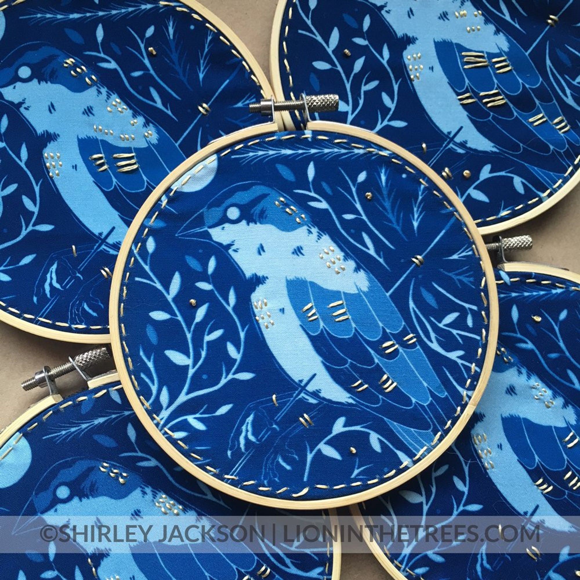 Blue cyanotype pieces done on fabric. The design features a House Sparry with an arrow piercing into it's front and out it's back. The bird is surrounded by flora and a moon in the upper left corner. 
There are extra, textural details done with a golden embroidery floss.