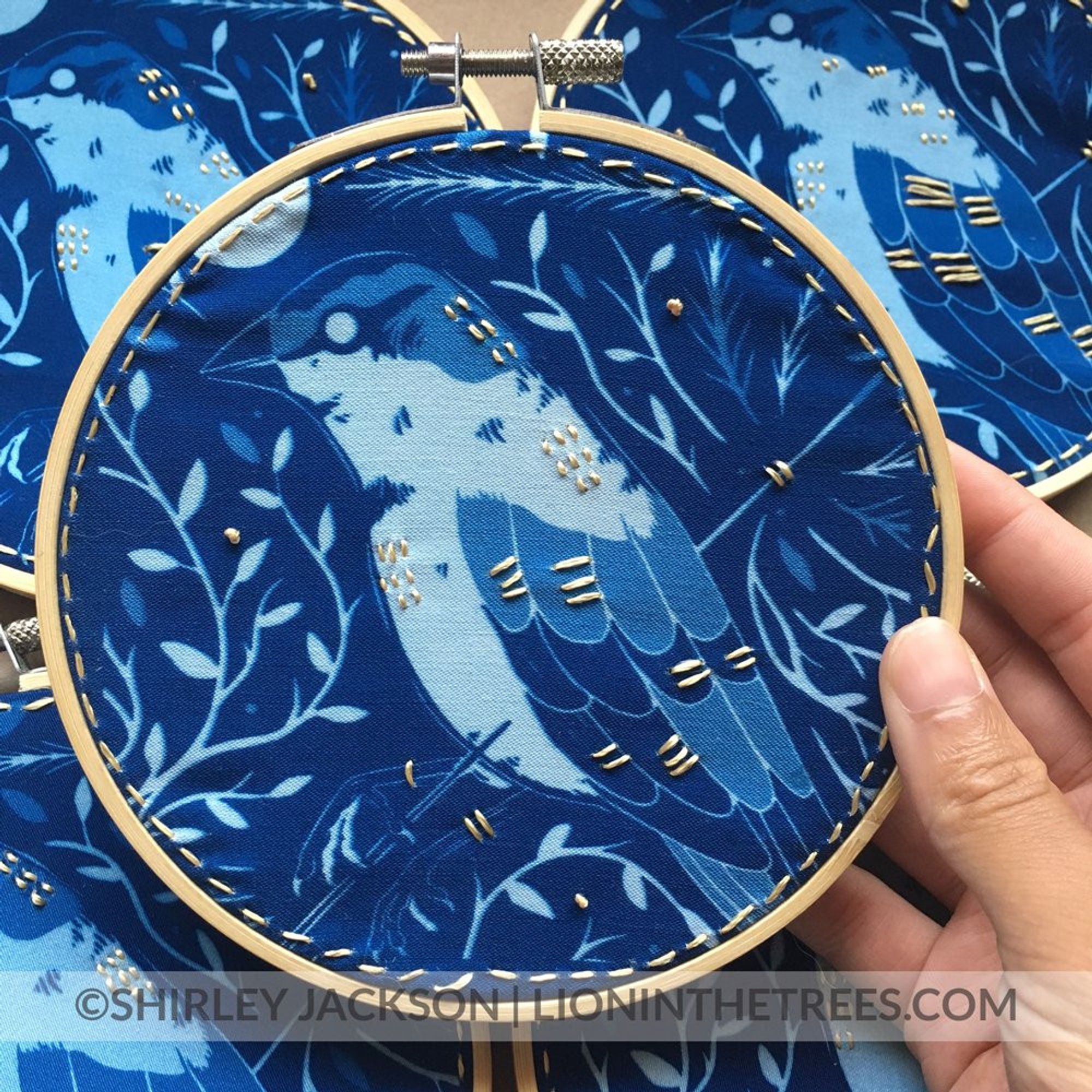 A more closeup photo of the blue cyanotype pieces done on fabric. The design features a House Sparry with an arrow piercing into it's front and out it's back. The bird is surrounded by flora and a moon in the upper left corner. 
There are extra, textural details done with a golden embroidery floss.