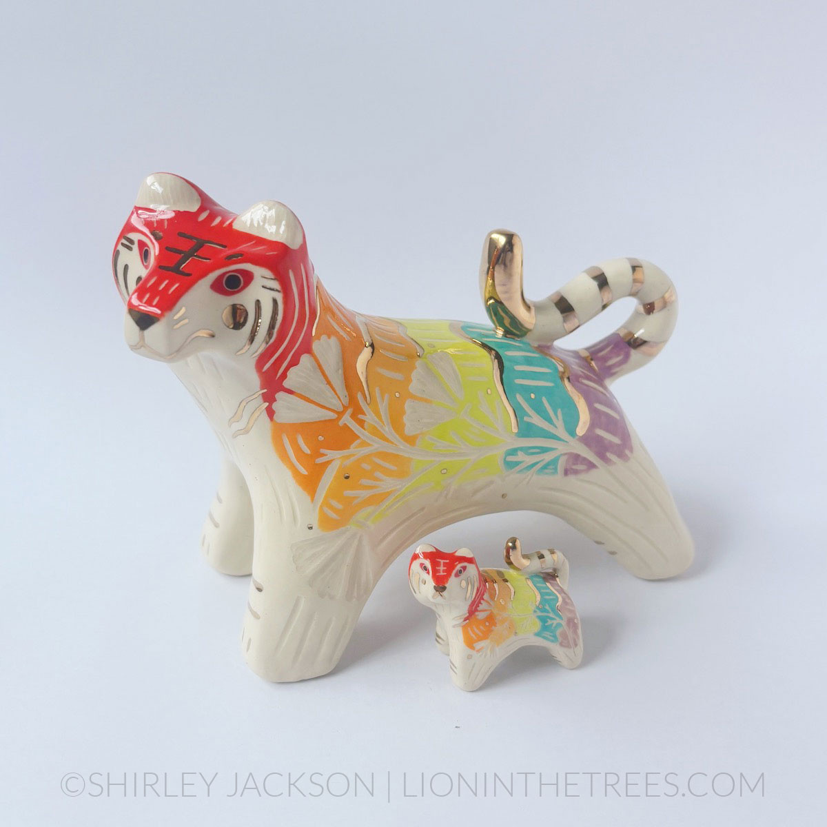 Two ceramic tiger sculptures done with an array of underglazes that follow a rainbow pattern with sgraffito details. One is a much larger version of the other.
