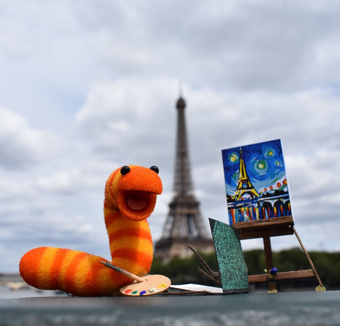 Slimey the worm is a character from Sesame Street. He is an orange and yellow striped worm and so small he can fit in the palm of your hand.
Here he is pictured plein air painting the Eiffel Tower because not only has he gone to the moon, but this worm also has artistic chops. Is there anything this worm CAN'T do?