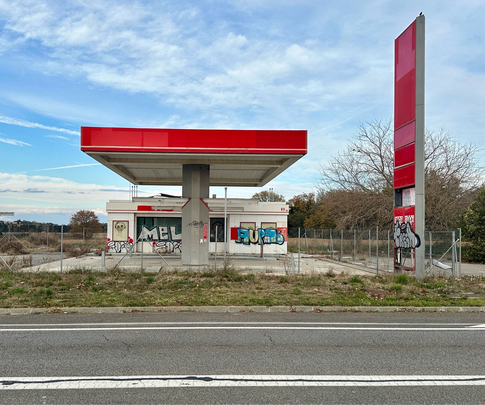 Verlassene Tankstelle, geschlossen und mit Graffiti besprüht. Landstraße , rote Dachumrandung und Preissäule, blauer Himnel leicht bewölkt