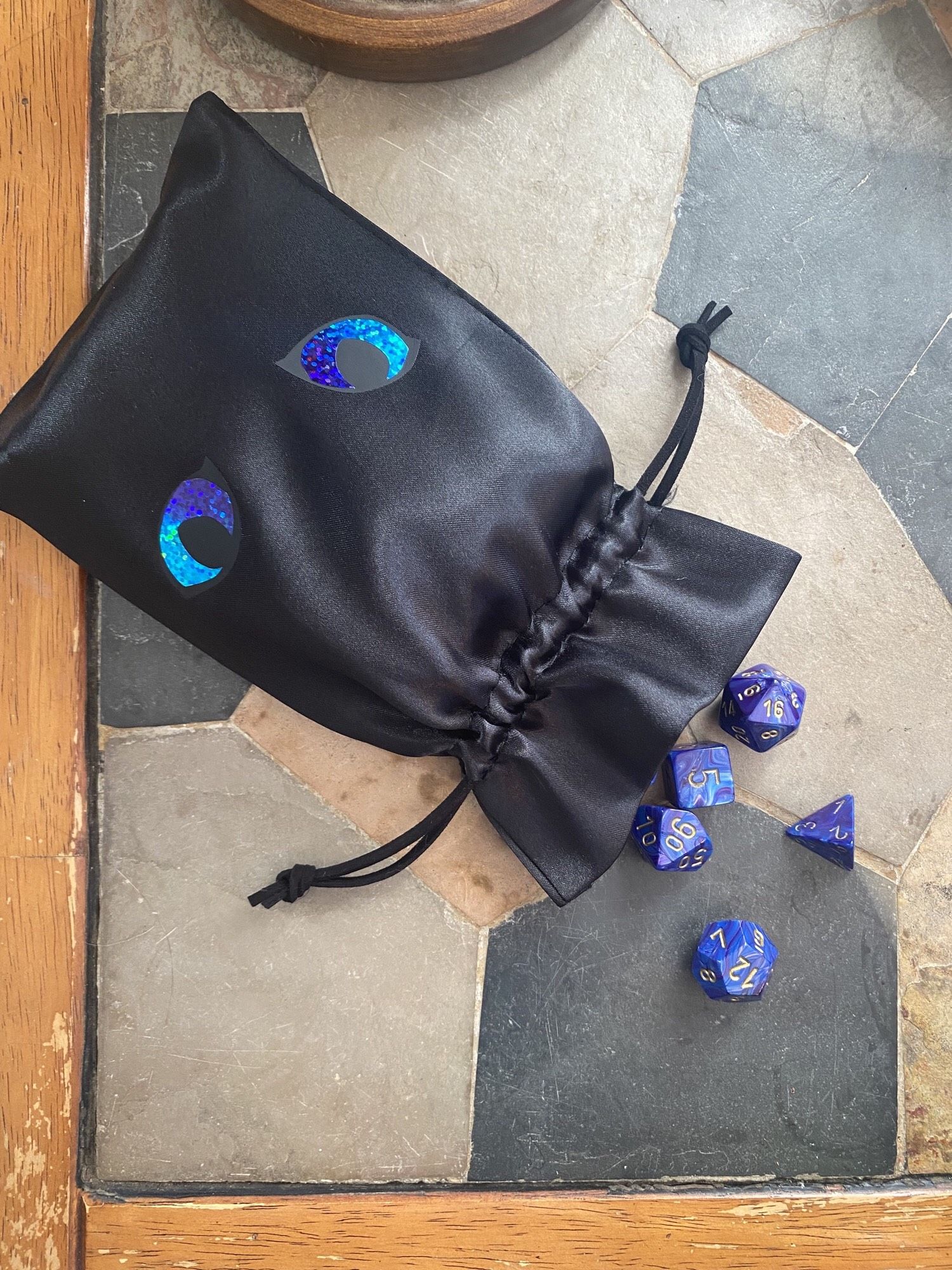 A shiny black satin drawstring bag laying open on a table with some blue dice spilling out. The bag has been embellished with matte black htv and holographic htv in the shape of cat’s eyes. The eyes are hypnotic and luminous blue and purple.