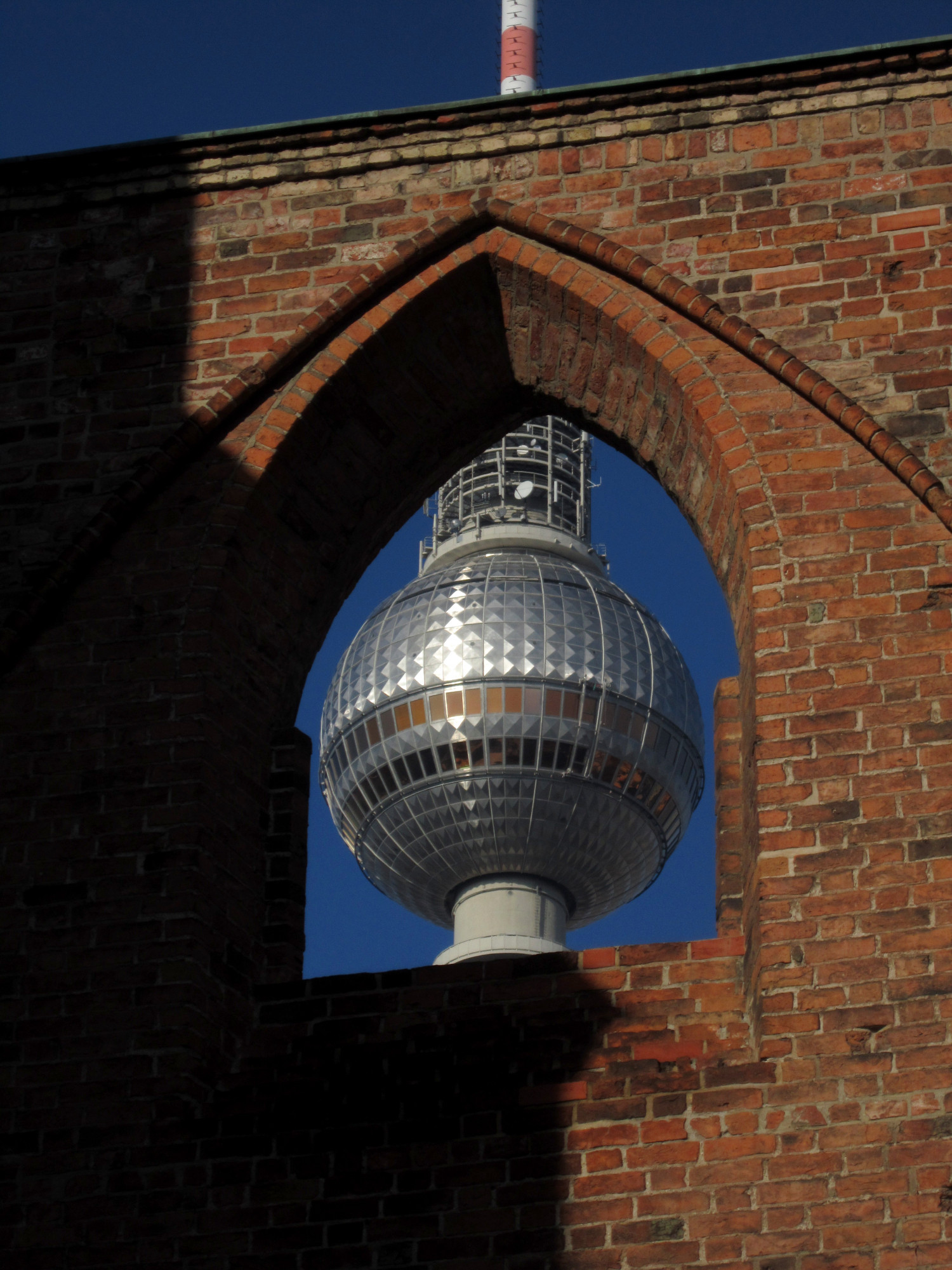 Eine Mauer aus rotem Backstein mit einer nach oben verjüngenden Öffnung. Dahinter sieht man die silberne Kugel des Fernsehturms vor wolkenlosem blauem Himmel.