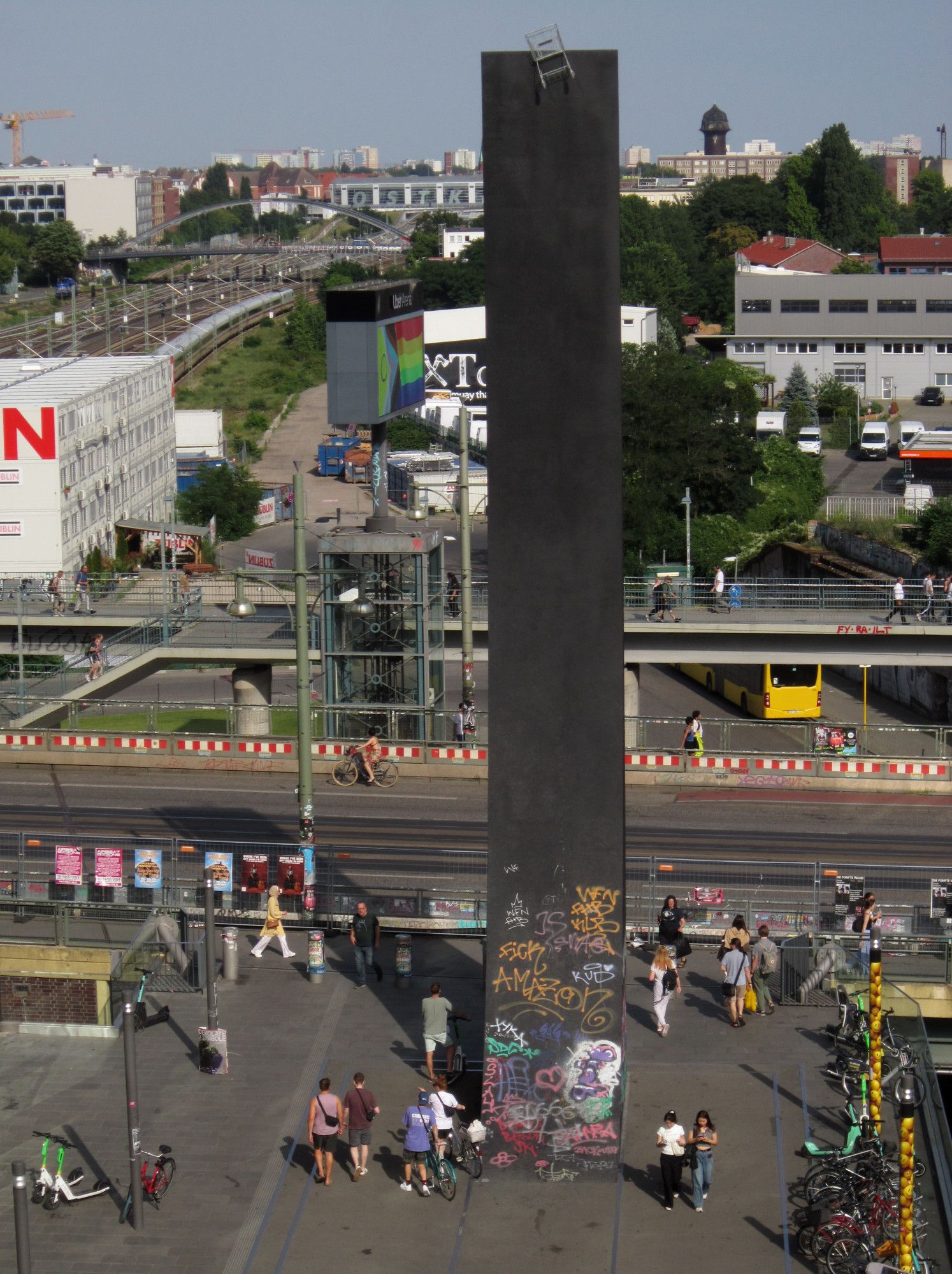 Blick auf eine Fußgängerkreuzung mit vielen sommerlich gekleideten Menschen. Mittendrin steht eine 23m hohe, 4m breite Wand aus dunklem Metall, auf der sich oben ein silberner Einkaufswagen befindet.