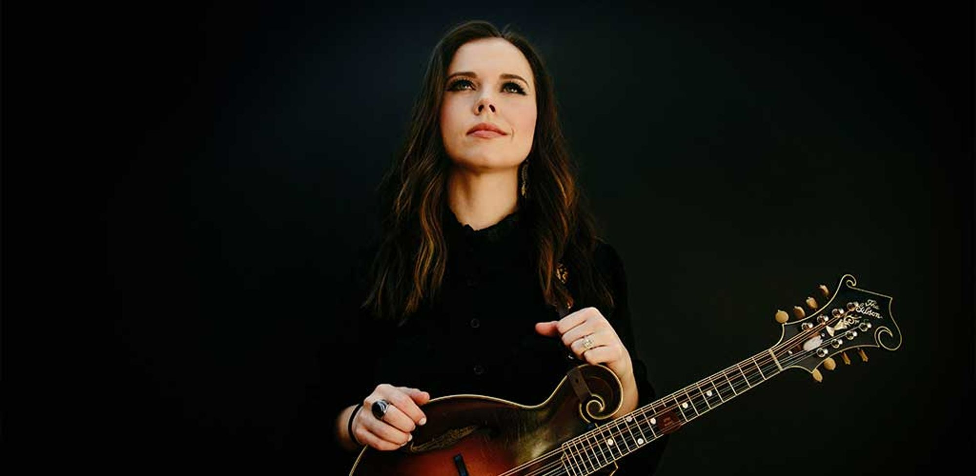 Sierra Hull holding her mandolin and looking upwards off camera. She's dressed in all black on a black background.