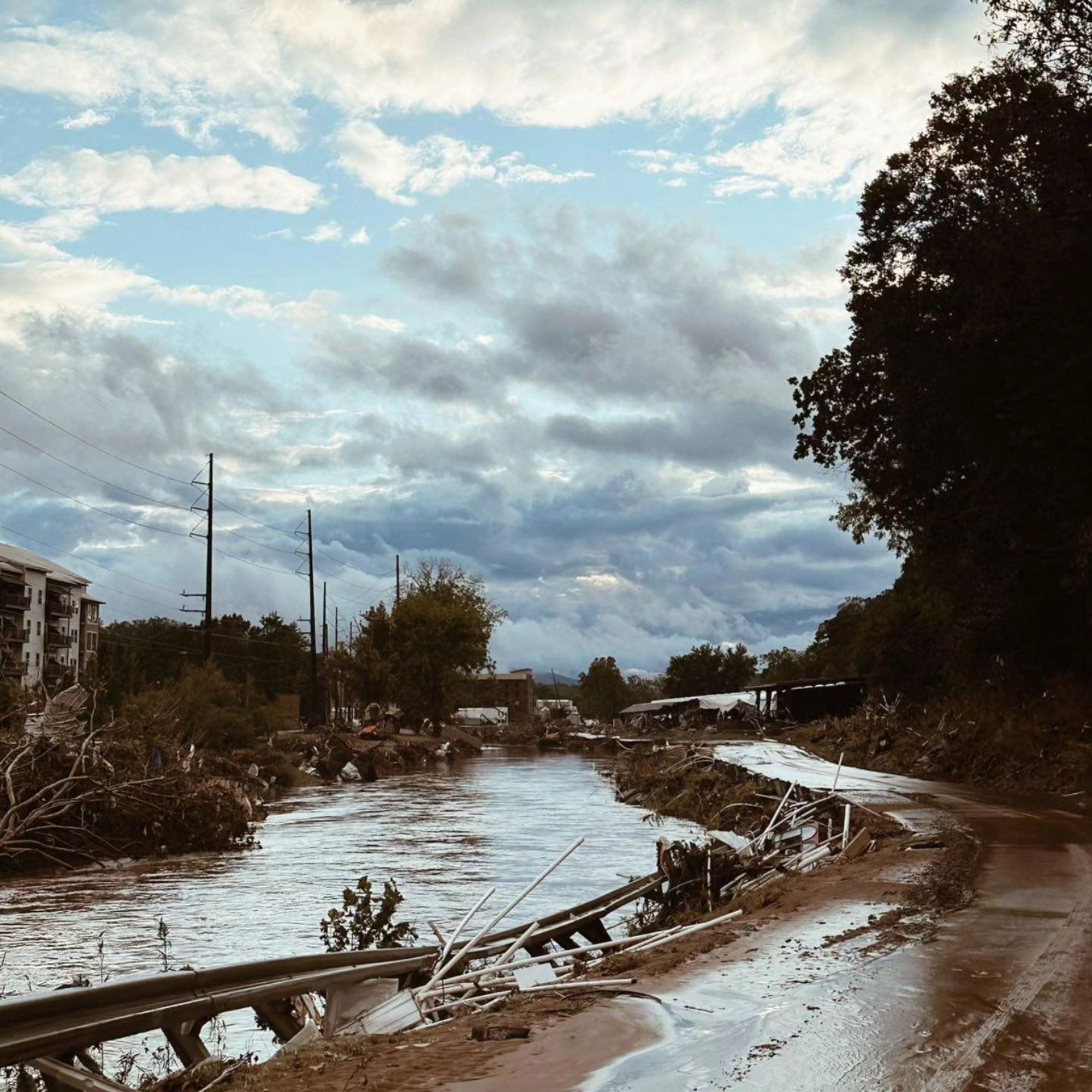 A photo of a washed out road in Asheville, NC. The signs of recently receded flood waters–including destruction of infrastructure and landscape–are everywhere, but the sky is blue with soft clouds.