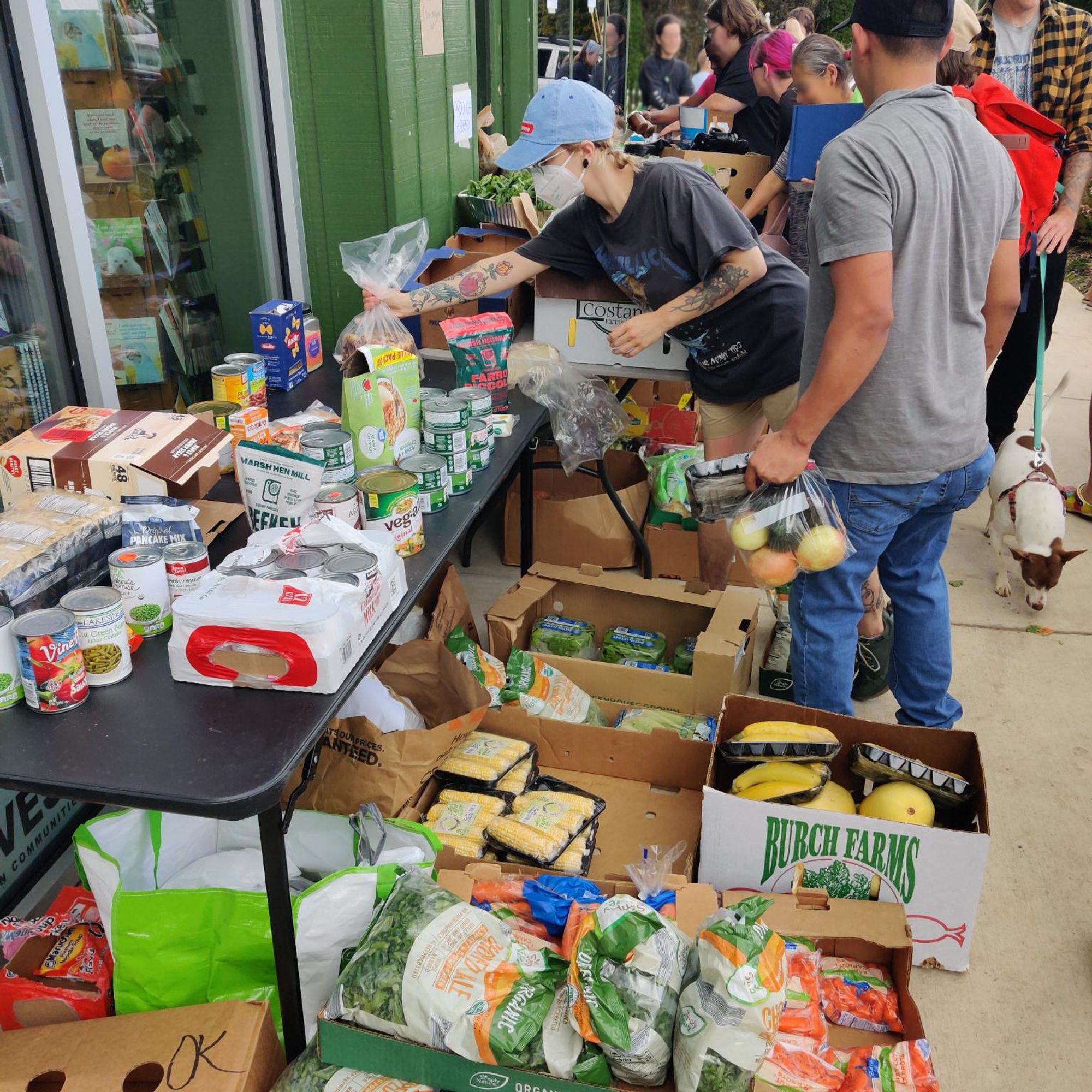 A photo taken outside Firestorm during the mutual aid food distro. A member of our collective moves food from boxes on the sidewalk to tables leaded with canned goods and produce while community members pick out things they need.