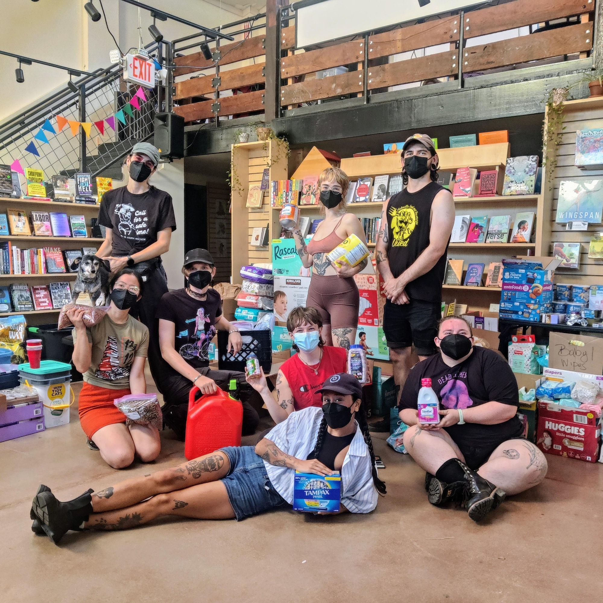 A photo of the distro volunteers at Firestorm Books on Sunday, October 6th. Eight masked humans and an elder dachshund pose for the camera with supplies in front of piles of diapers, pet food, baby supplies, and other items. Bookcases have been pushed aside but are visible in the background.