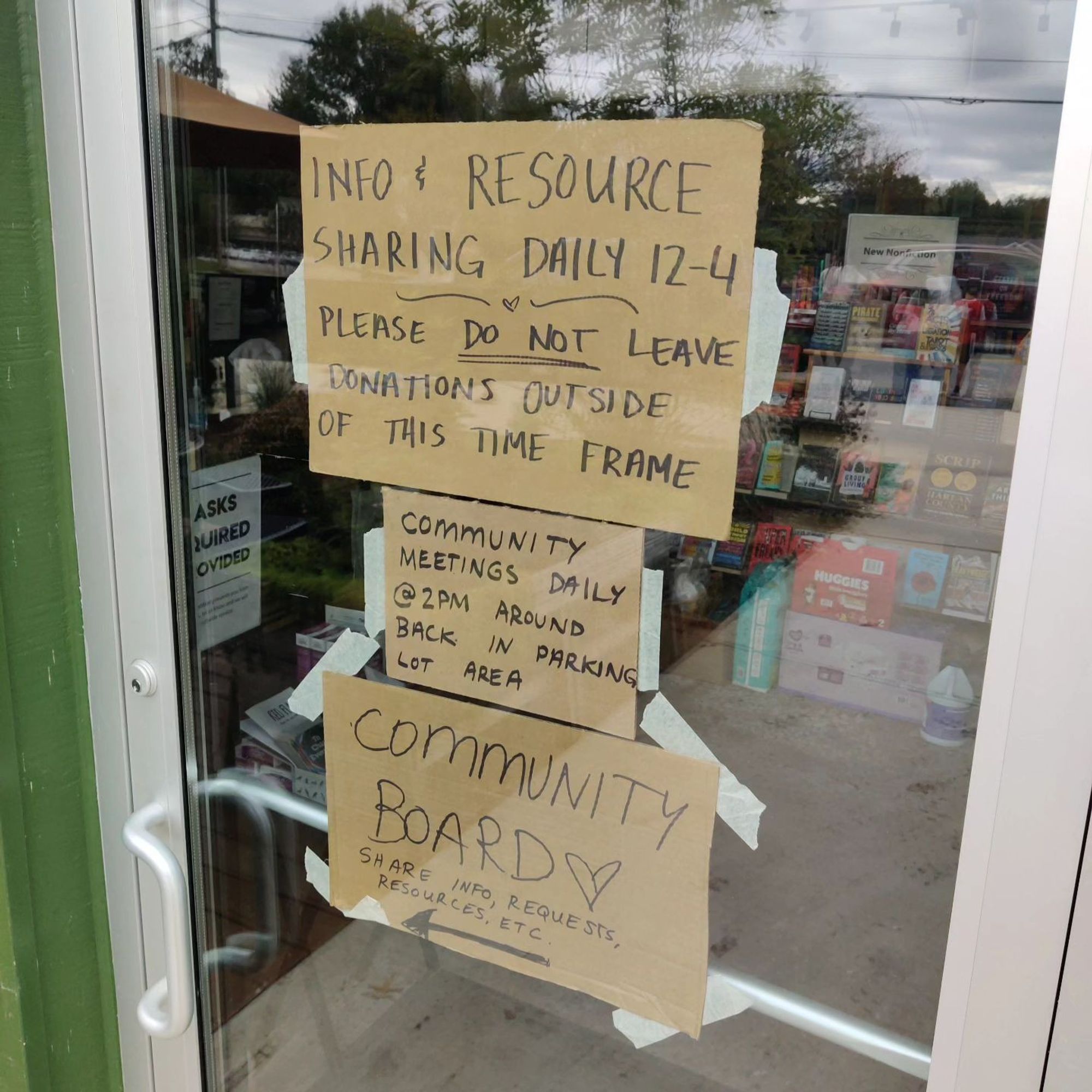 A photo of Firestorm Books' front door. Cardboard signs taped to the glass read: "Info & Resource Sharing, Daily 12-4. Please do not leave donations outside of this time frame. Community meetings @ 2pm around back in parking lot area. Community Board: Share info, requests, resources, etc." Inside the building piles of supplies are visible.