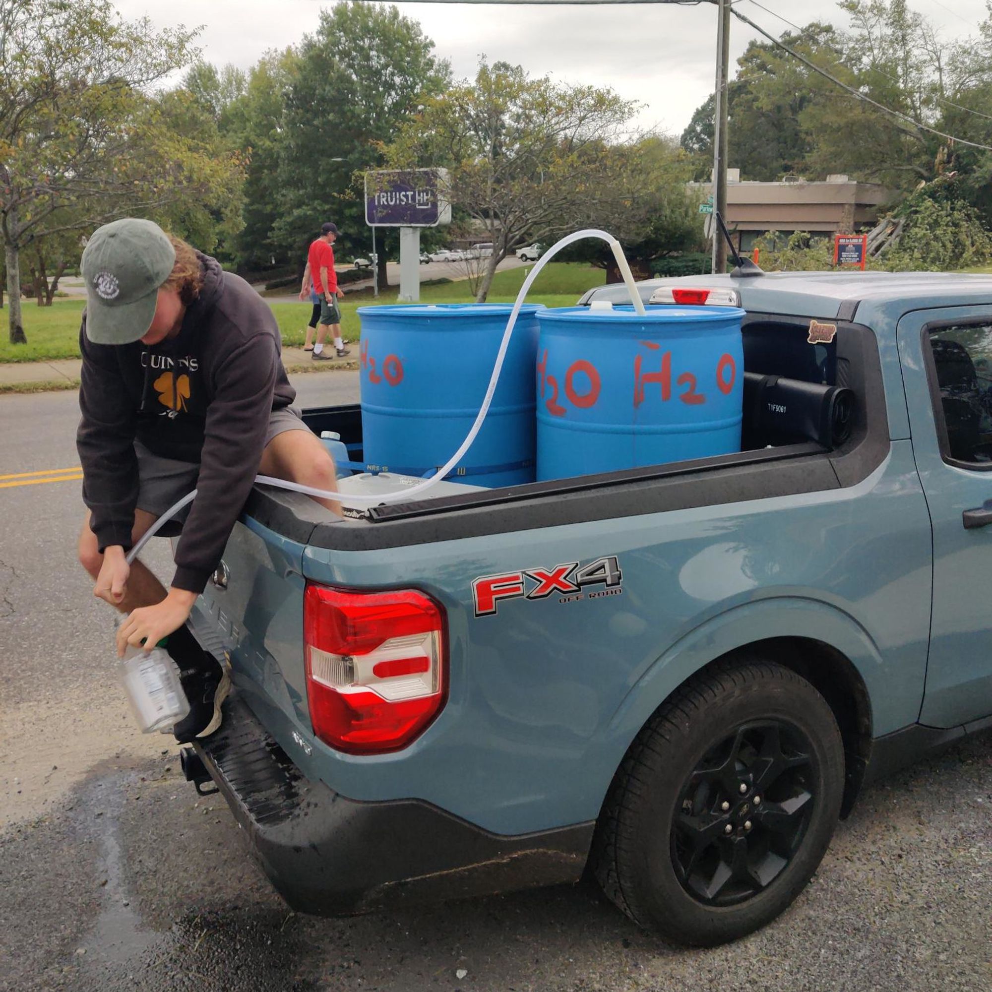 A photo of an individual in the back of a truck siphoning water into a glass bottle. The water is coming from one of two small barrels labeled “H2O.” In the background, a downed tree is visible.