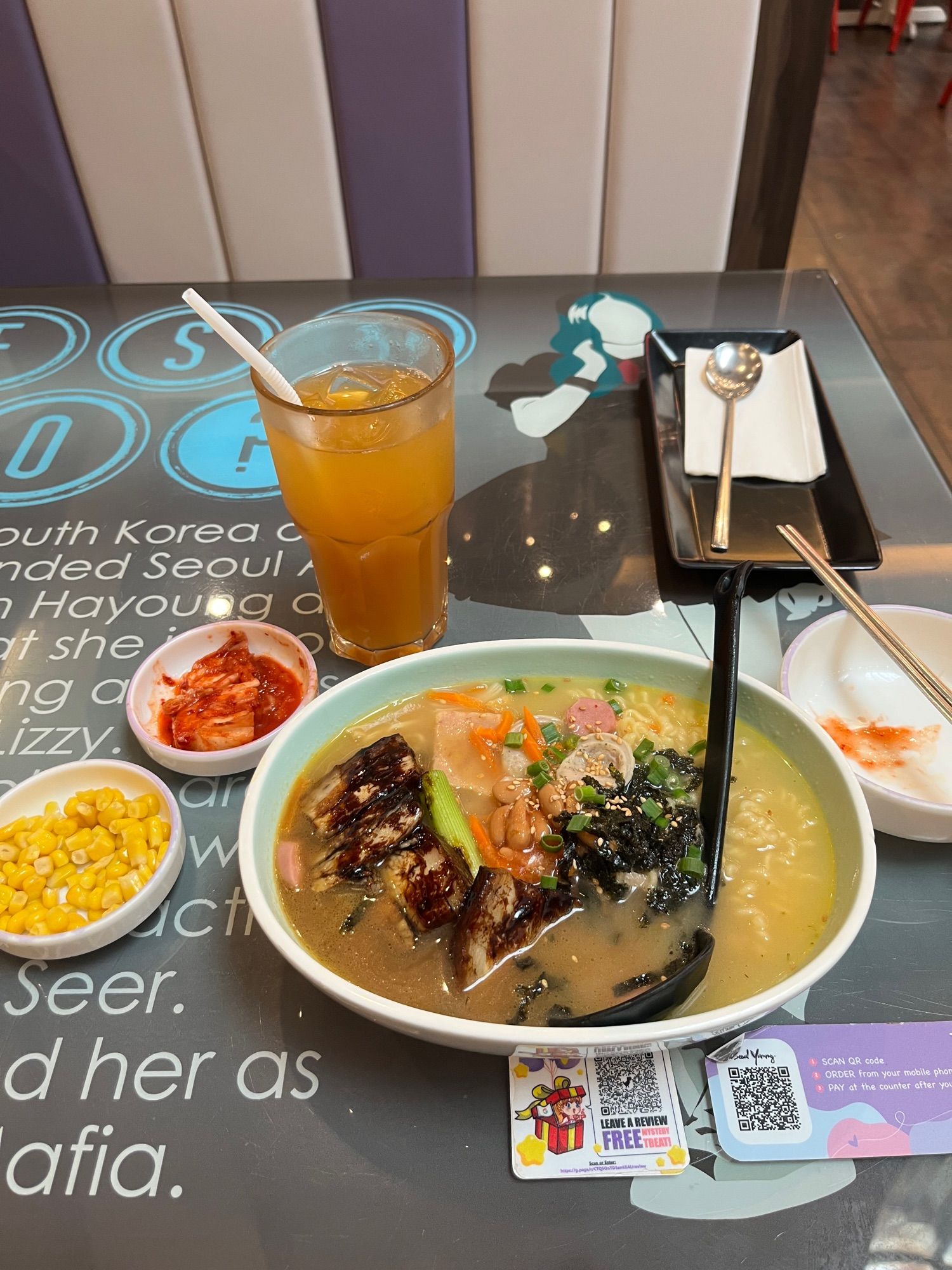 A large bowl of clear stew with bits of seaweed, spring onion, ham, luncheon meat, sliced sausage, and instant korean noodles. There’s a glass of ice lemon tea, and smaller plates with boiled sweet corn, and kimchi on the side.