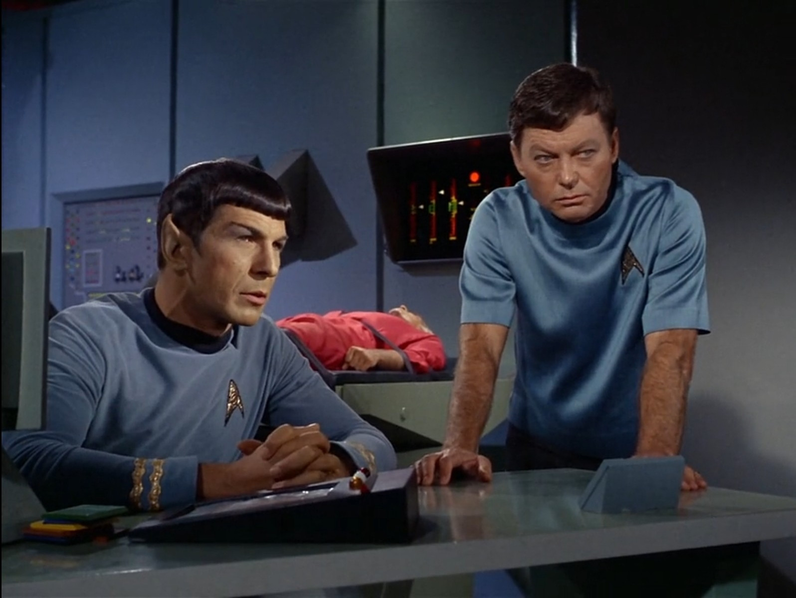 spock and doctor mccoy from star trek the original series in sickbay. they're sitting and standing by the desk, respectively, and there's a patient lying on a bed behind them. they're both looking ahead.