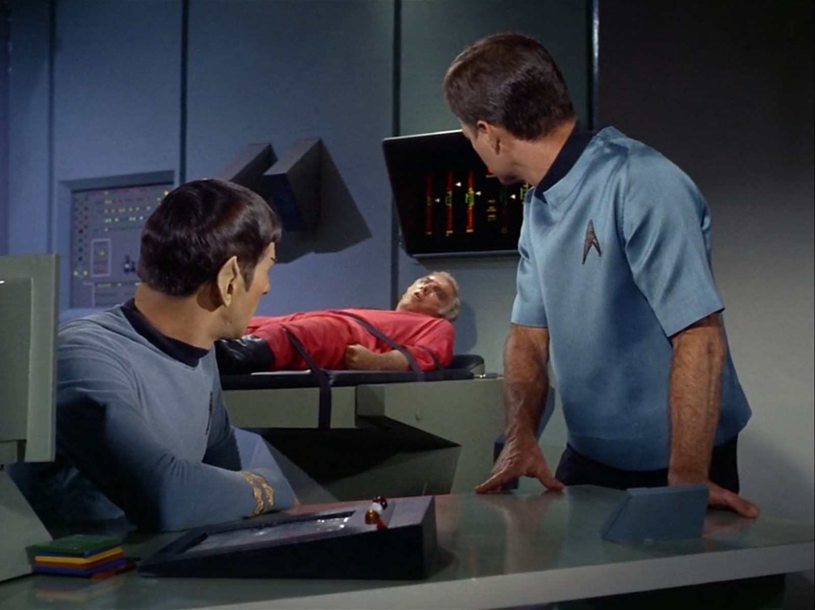 spock and doctor mccoy from star trek the original series in sickbay. they're sitting and standing by the desk, respectively, and there's a patient lying on a bed behind them, now awake. spock and bones are looking at the patient.