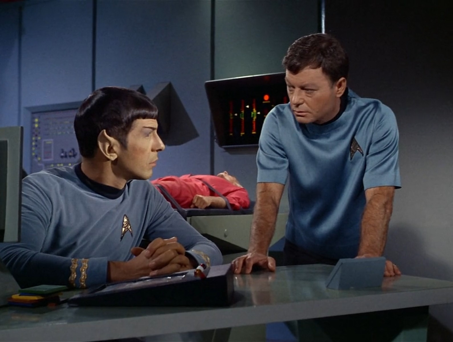 spock and doctor mccoy from star trek the original series in sickbay. they're sitting and standing by the desk, respectively, and there's a patient lying on a bed behind them. they're looking at each other.