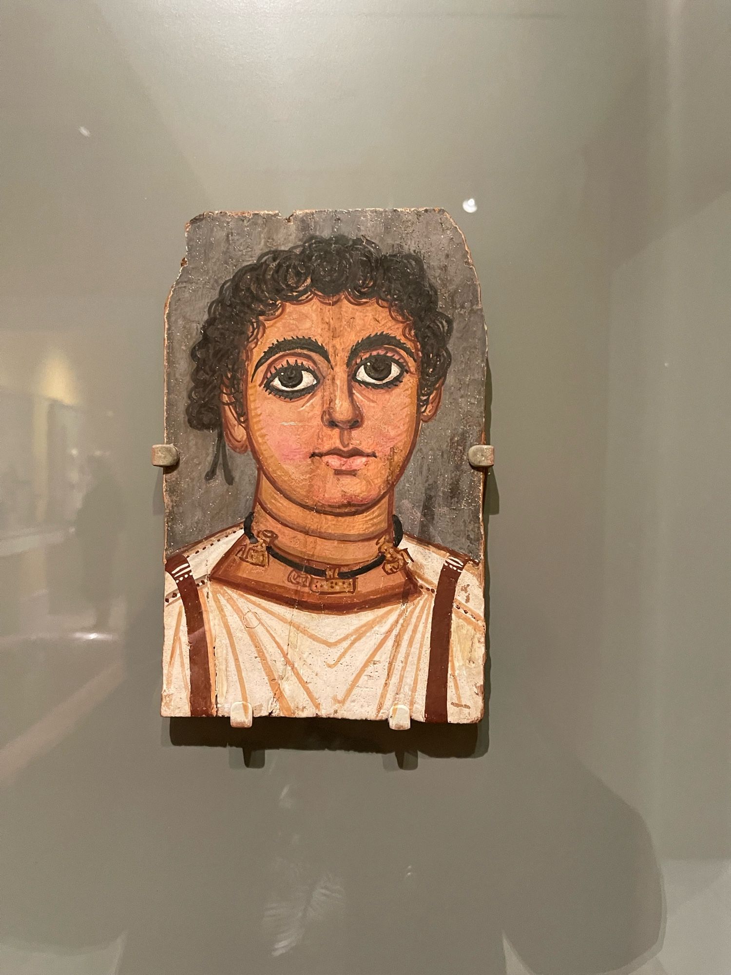 Beginning in the 1st century CE, a realistic portrait of the deceased - painted on a wooden panel—might be placed on the outer wrappings of a mummy. The big eyes and bushy eyebrows seen in this work are typical stylistic traits from an area of northern Egypt called Fayum. While the boy's clothing was common for the period, his necklace with three amulets is rare. The two outer figures represent the divine couple Isis and Serapis, and between them is a metal leaf, probably a passport for the dead. These amulets were often inscribed with instructions on how to behave in the afterlife.