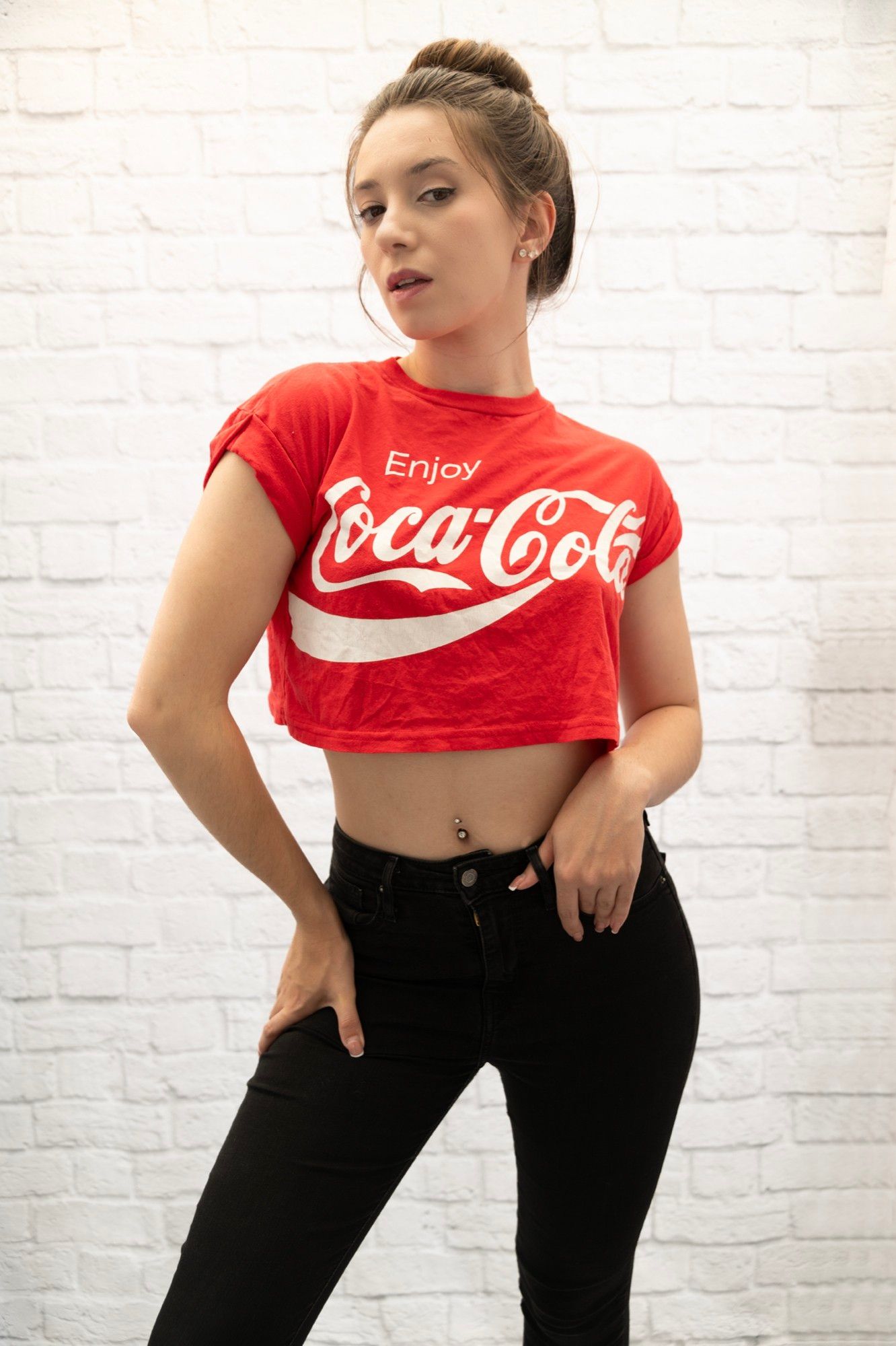 A studio-style photo of a dark-haired young woman posing. She's wearing black trousers and a short, red "CocaCola" tee-shirt.