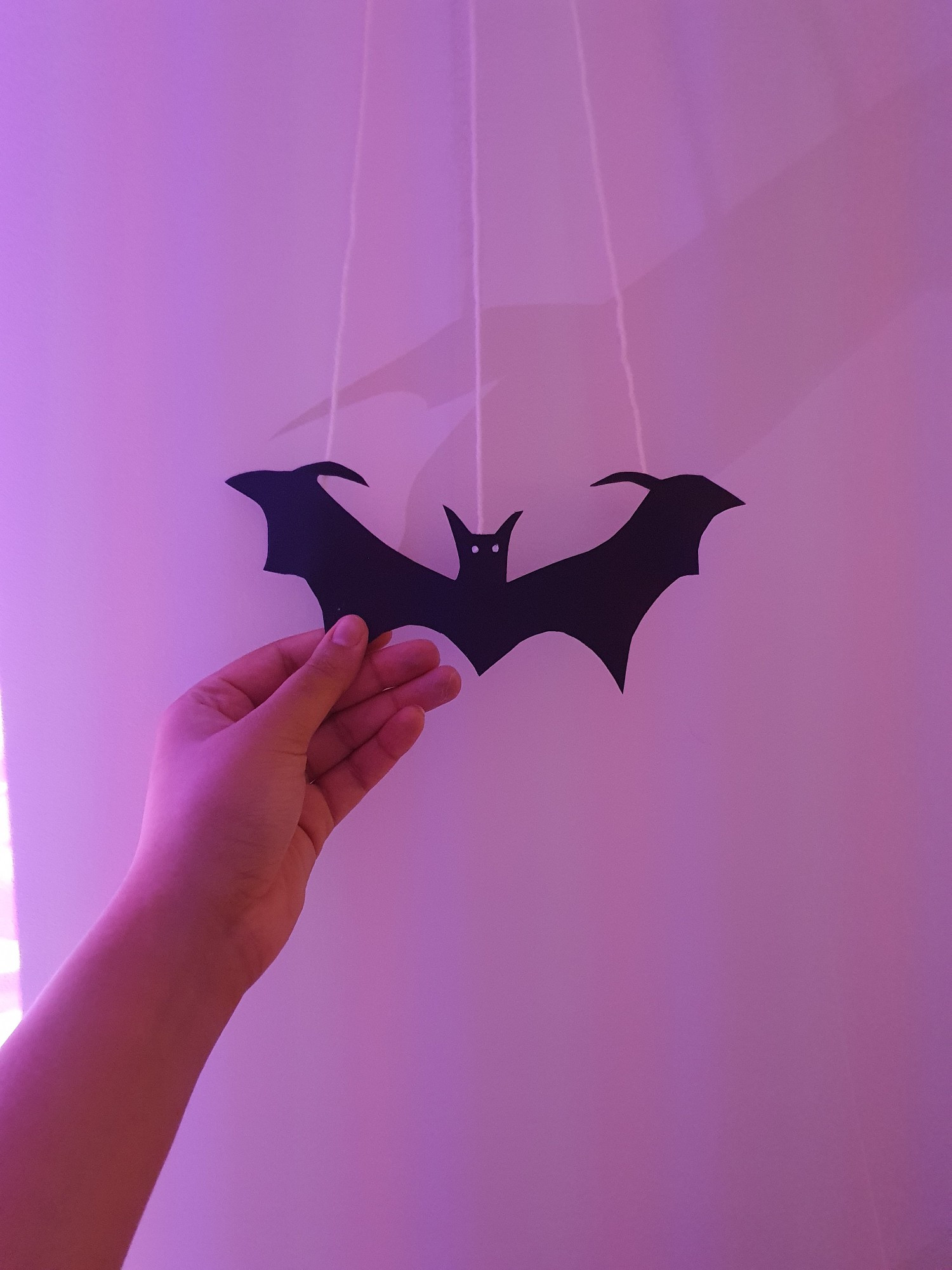 Me holding a paper black bat I cut out, hand outstretched, the paper bat is attached to the wall via three strands of white yarn