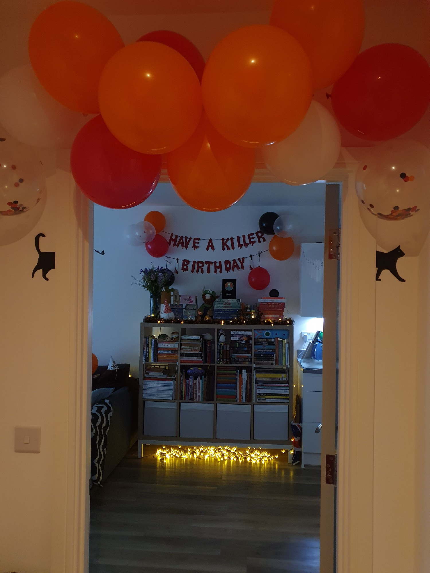 orange red white and confetti balloon garland over door frame, with a black cat split and stuck on both sides so it's 'passong through', with 'have a killer birthday' on the wall in the background surrounded by more balloons and a bookcase below covered in fairy lights