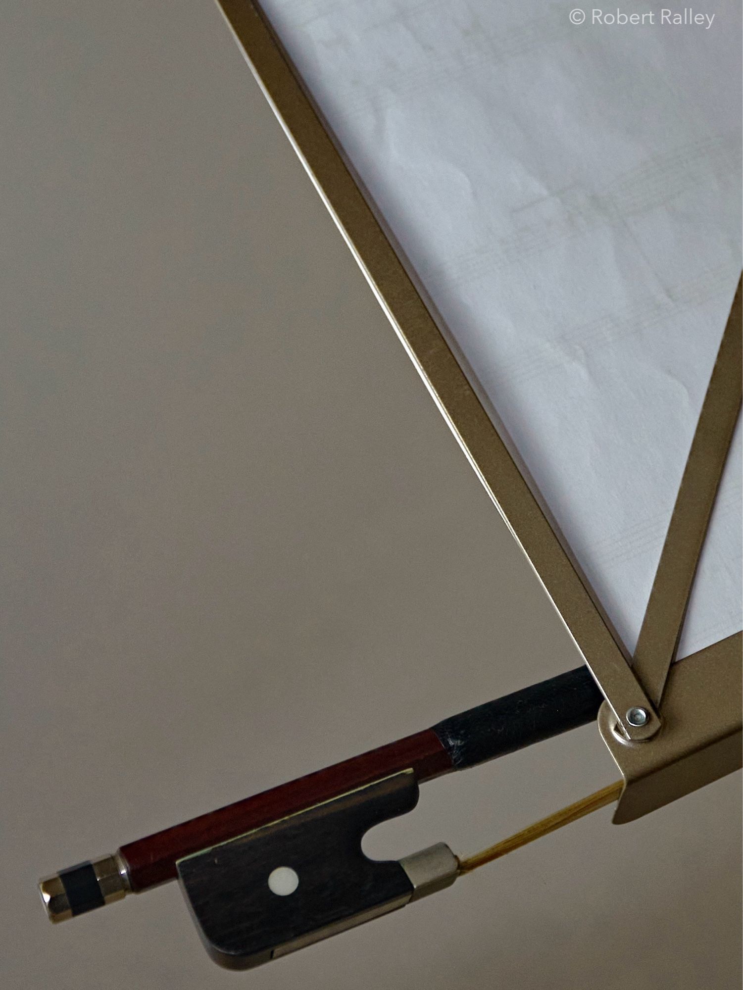 A view upwards from below a lightweight music stand with sheets of music on it, showing just the bottom corner of the sheets and metal frame. Sticking out from it is the end of a cello bow, resting on the music shelf.