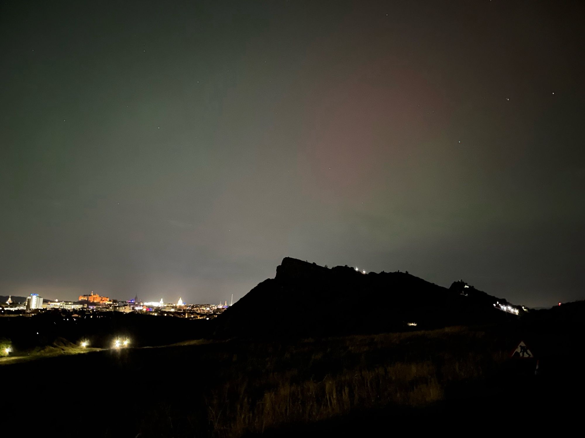 The sky still pale green into black, with a faint oval of red hanging over the distinctive squat triangular shape of Salisbury Crags. The crags are speckled with the headlamps of walkers. To the left in the distance, the city of Edinburgh is a strip of illumination with the castle at its centre.