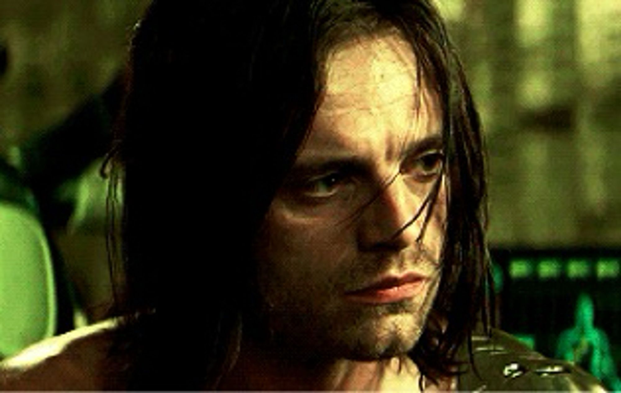 Sebastian Stan as Bucky Barnes, the Winter Soldier. His head is tilted slightly to one side, has mouth flat, his eyes distant and sad. His lank almost shoulder length hair frames his face.