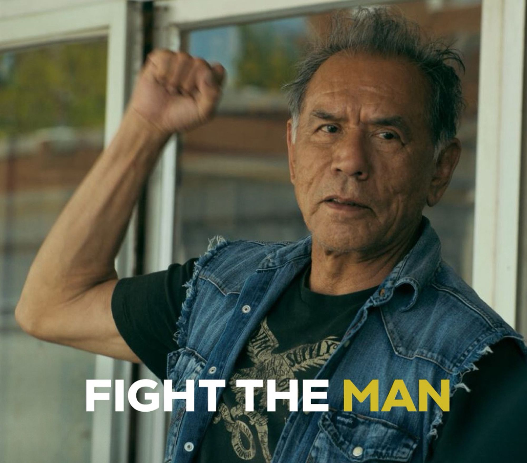 Wes Studi as Bucky, one of the elders in Reservation Dogs. His hair is gray and short, his right fist raised high, his strong features lined with argue. He is dressed in a black t-shirt with a sleeveless denim jacket. The text on the picture reads Fight The Man.