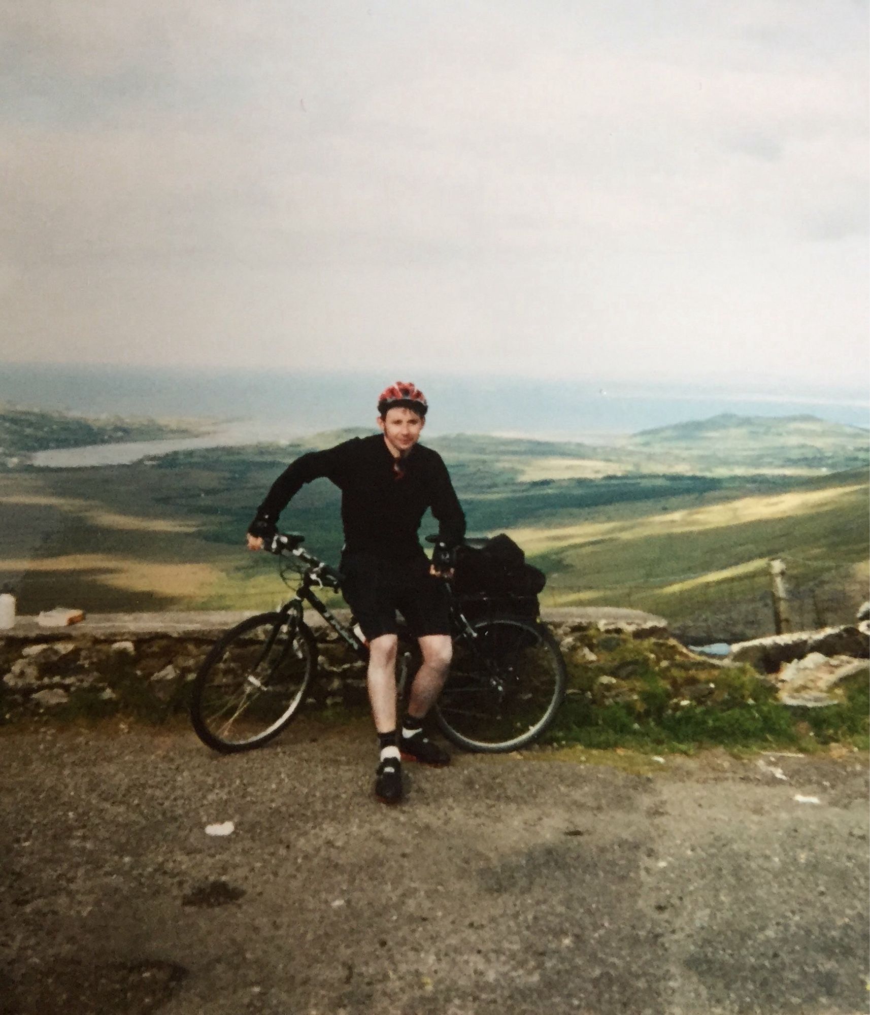 A picture of a cyclist in his twenties taken at the top of Connor Pass in Kerry. He is dressed in a black long sleeved  cycling base layer with black cycling kits and baggy black off-road cycling shorts, and black cycling shoes. On his head is a dark red cycling helmet. He leans against a dark green mountain bike which is propped up against a low stone wall. A single rear pannier is attached to the bike’s rear luggage rack. Behind him the land slopes down to the Atlantic Sea.