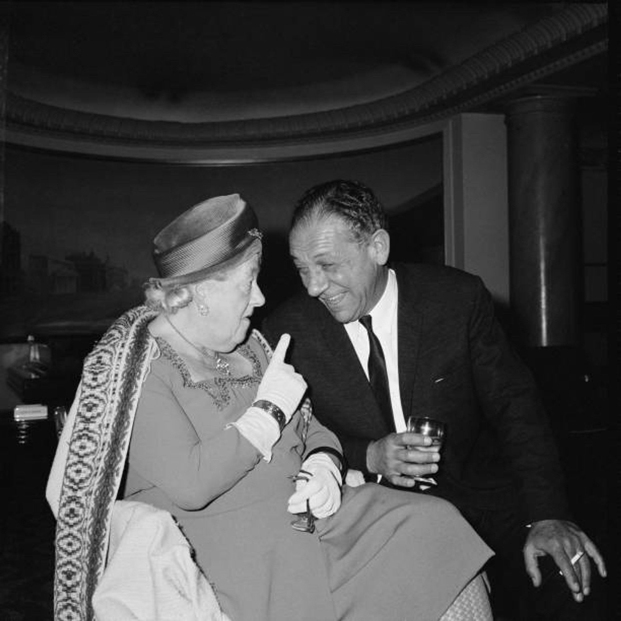 Margaret Rutherford and Sid James sat together in theatre seats in a black and white photo.