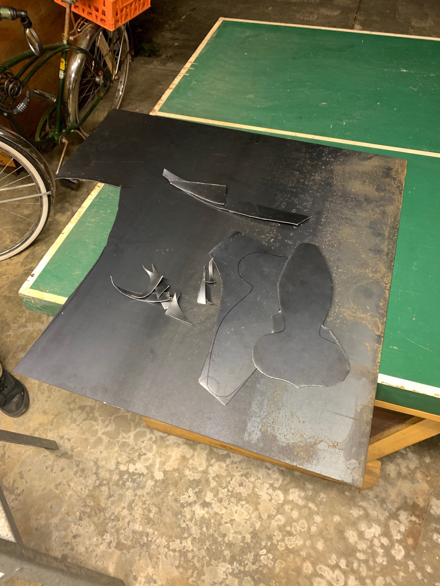 flat kneecop pieces cut out in angular pieces from a large sheet of metal, with one almost entirely cut out; the scraps from cutting are placed next to the pieces