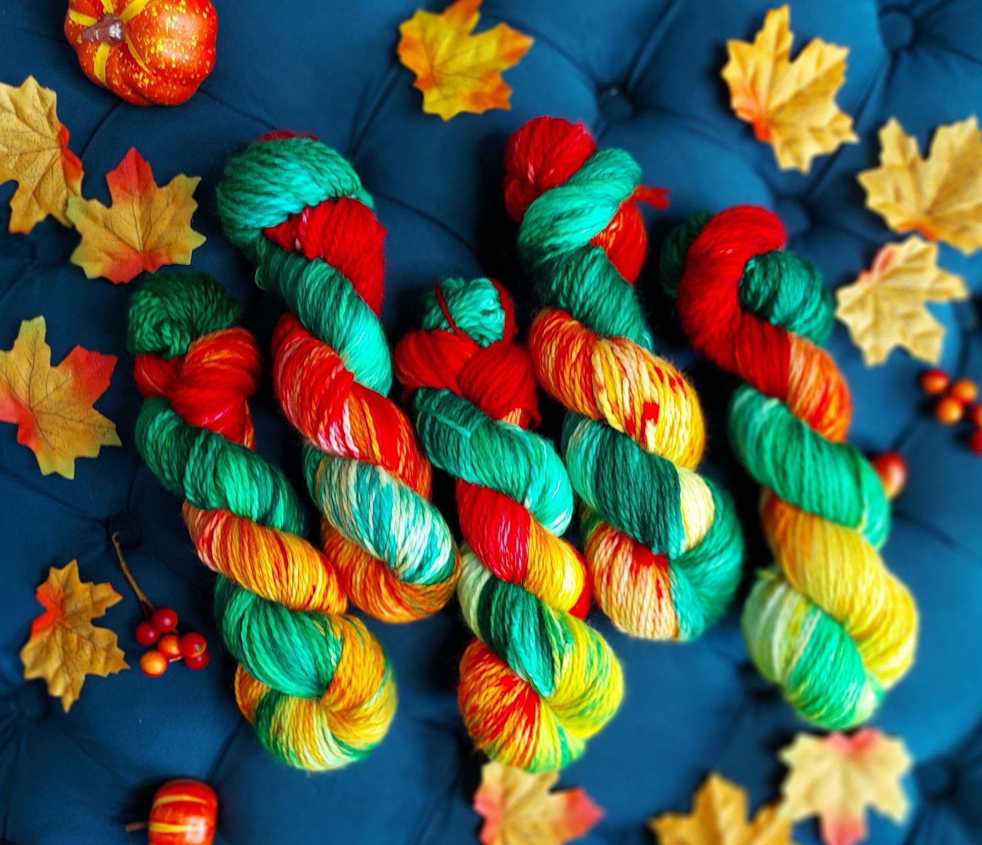 5 skeins of variegated yarn. The colorway is the colors of fall leaves transitioning to orange, red and yellow. There are leaves around the skeins with acorns and a small squash in the upper left.