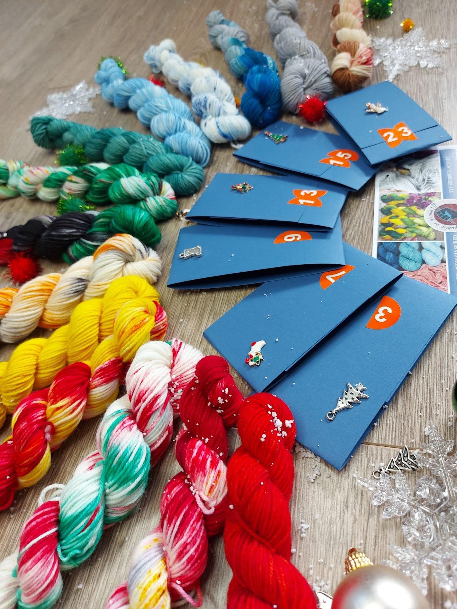 Angled shot of a rainbow line up of 14 holiday themed mini skeins. Surrounded by tree ornaments and snowflakes, 6 folded cardboard facts and a postcard.