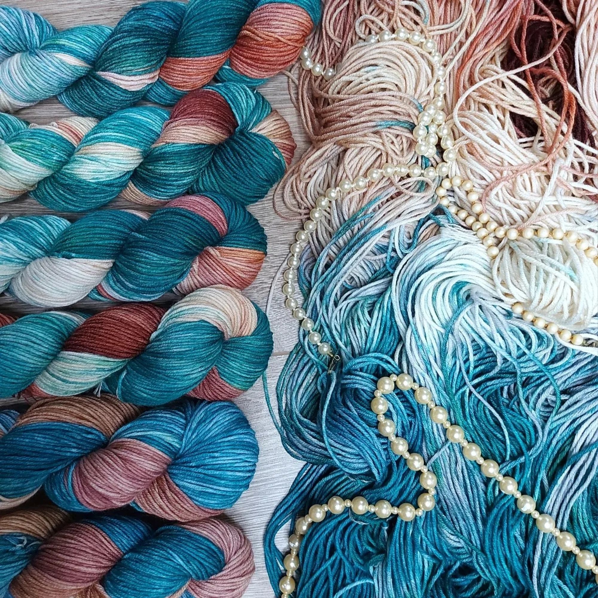 skeins of mystic magi teal with one open skein on the right. There are pearl necklaces scattered