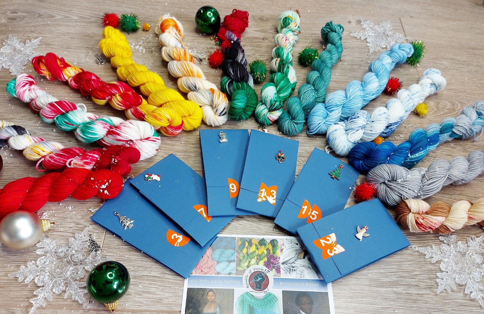 a rainbow line up of 14 holiday themed mini skeins. Surrounded by tree ornaments and snowflakes, 6 folded cardboard facts and a postcard.