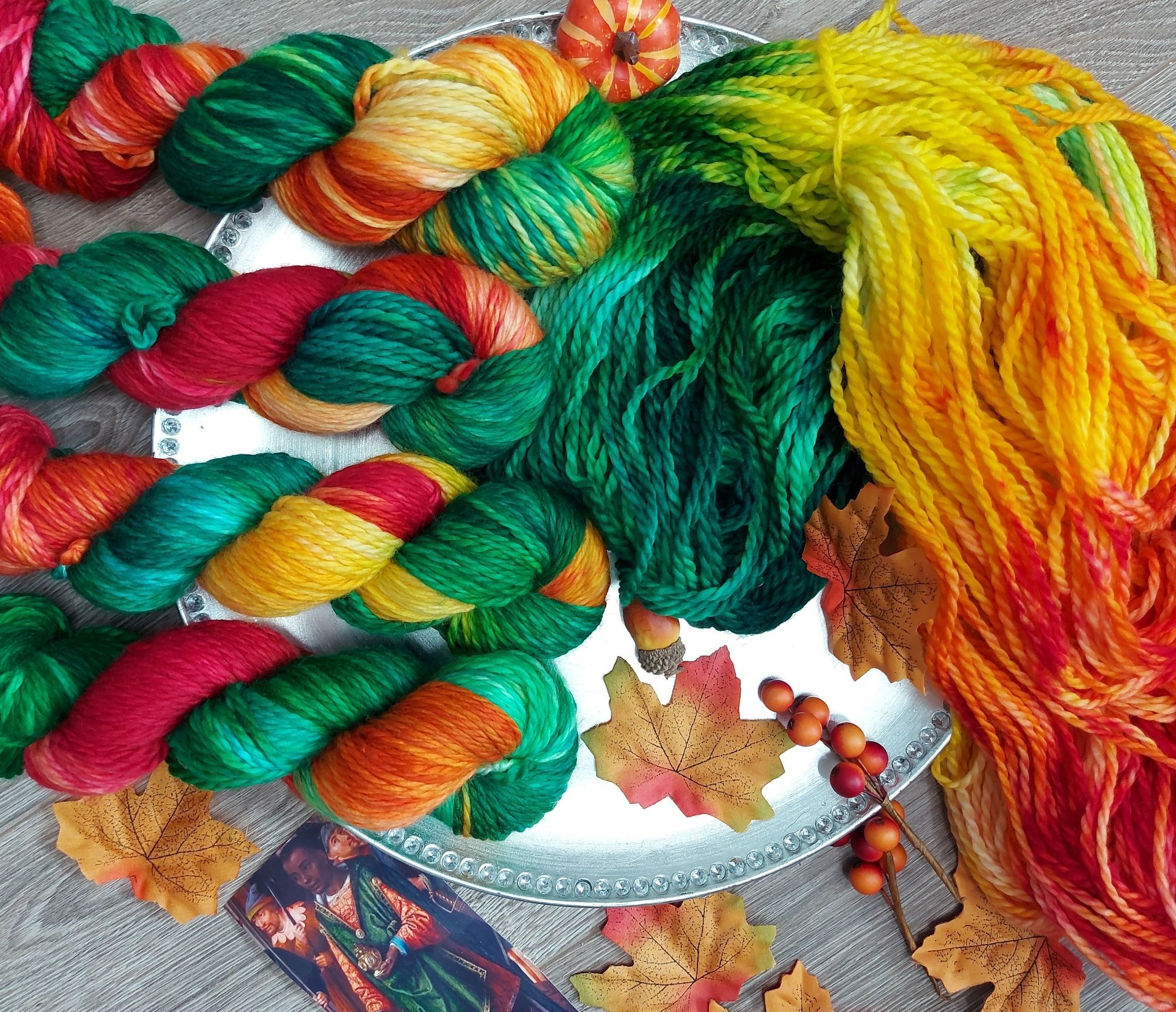 Red, yellow, and green yarn on a silver platter, adorned with Autumn leaves and berries. A photo of a magus dressed in the same colors is also in the shot.