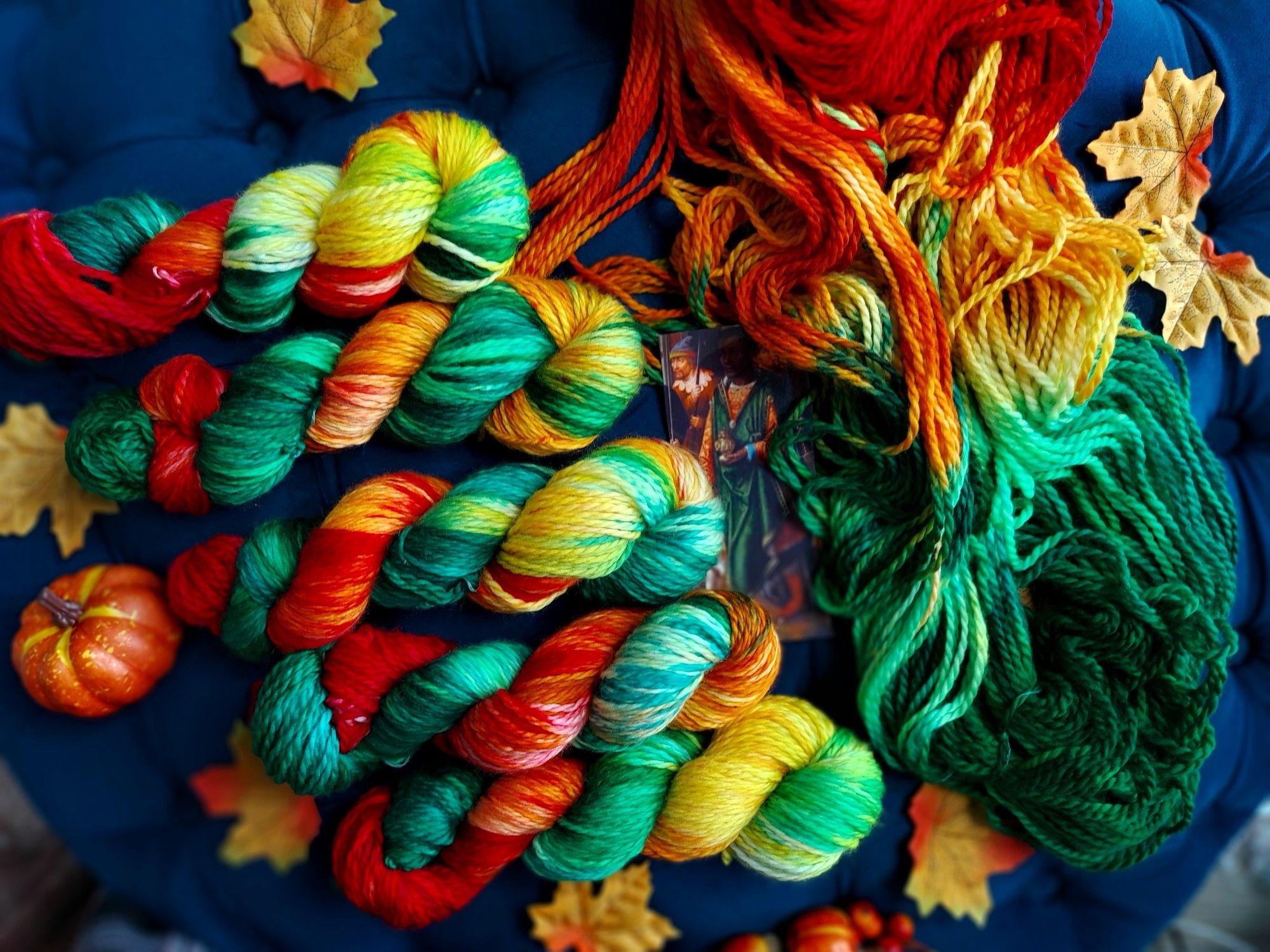 Skeins of red, yellow, and green yarn lined up, and decorated with with Autumn leaves, berries, and a miniature pumpkin.