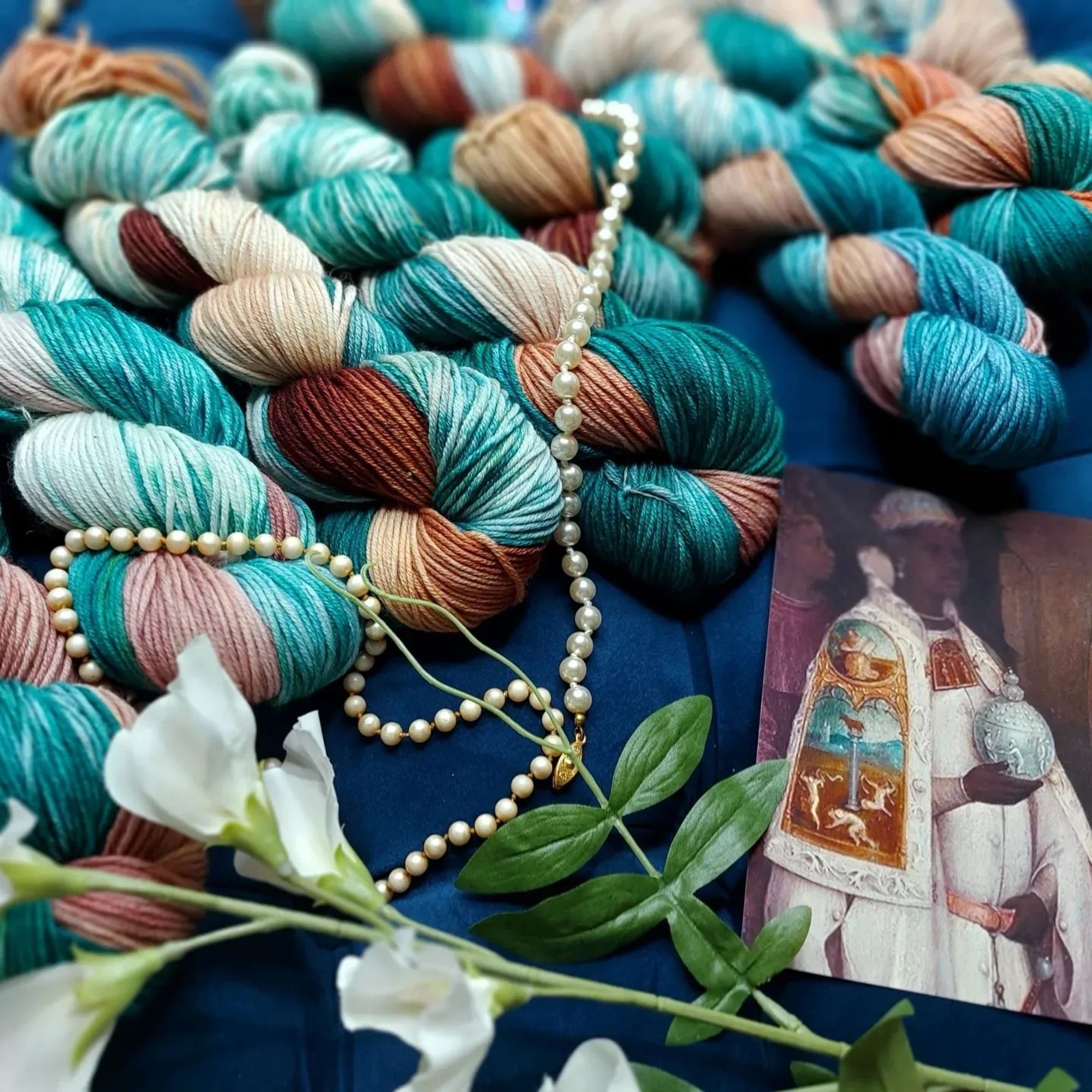 Angled shot of mystic magi teal with pearls, leaves and an image of a 16thc Black figure in a white teal and brown robe holding a light blue globe.