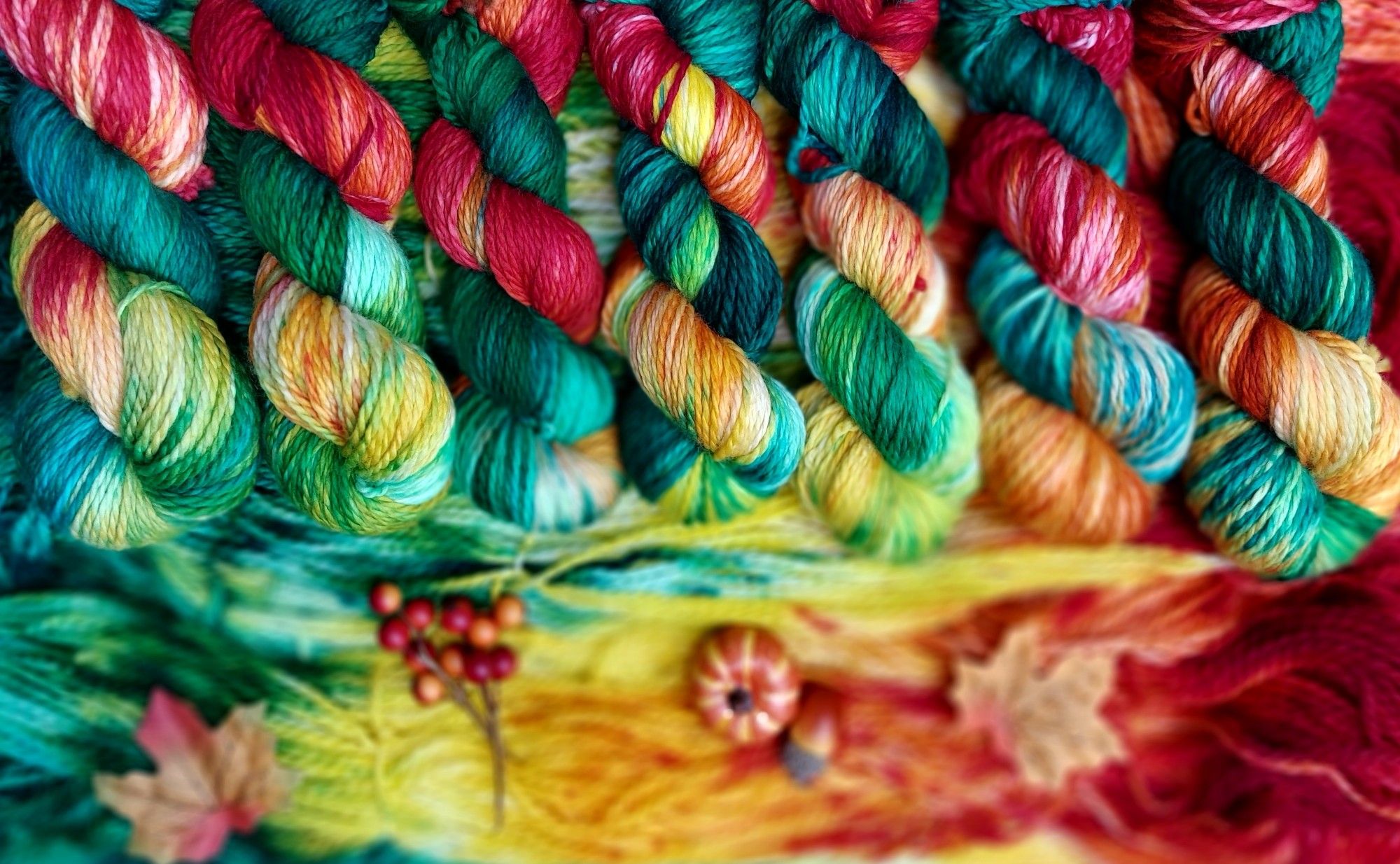 Skeins of red, yellow, and green yarn lined up, and decorated with with Autumn leaves, berries, and a miniature pumpkin.