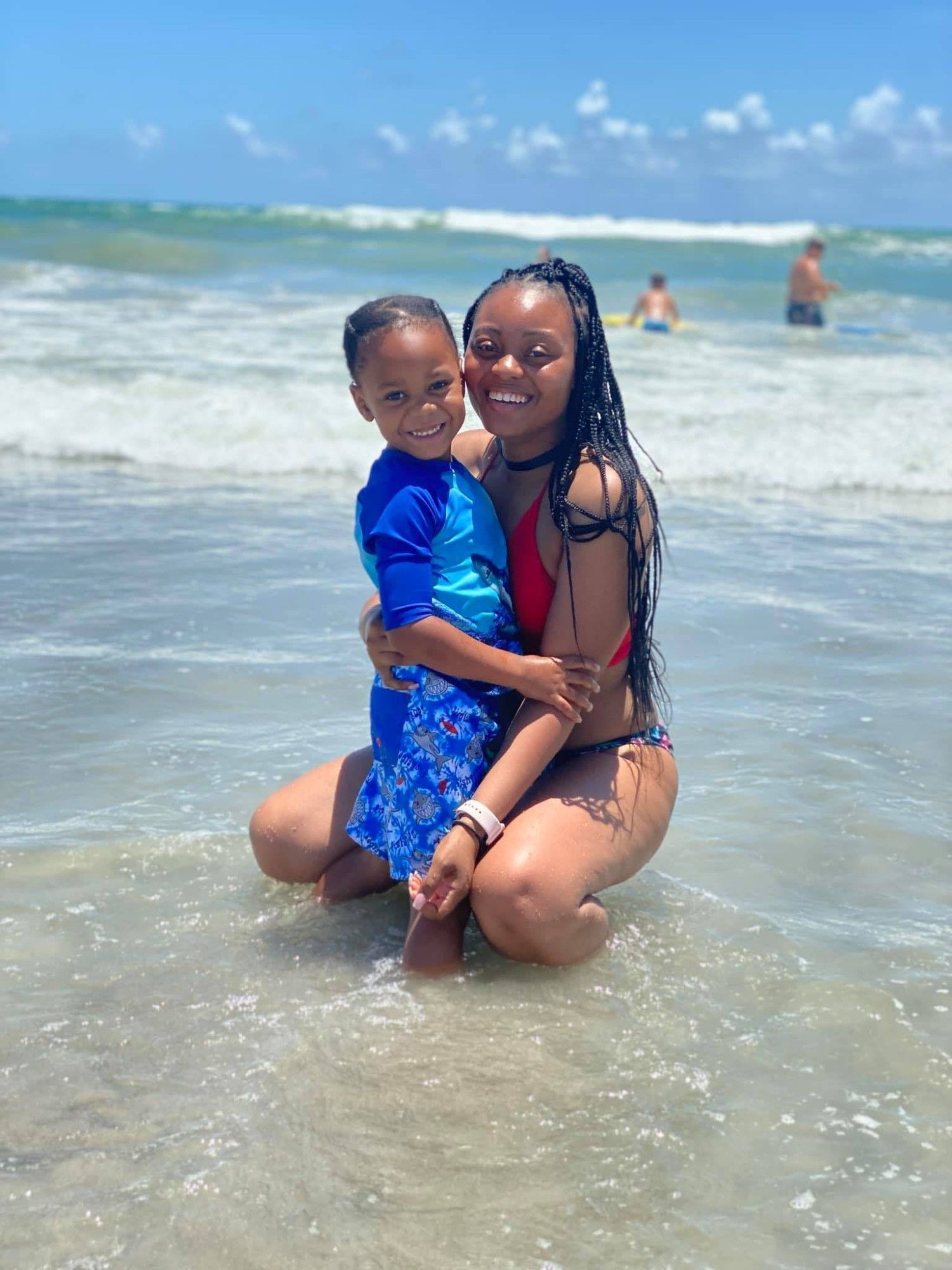 A photo of Amber Thurman and her son smiling for the camera at a beach. Amber is crouching down while hugging her son close. The two are both dressed in swimwear.