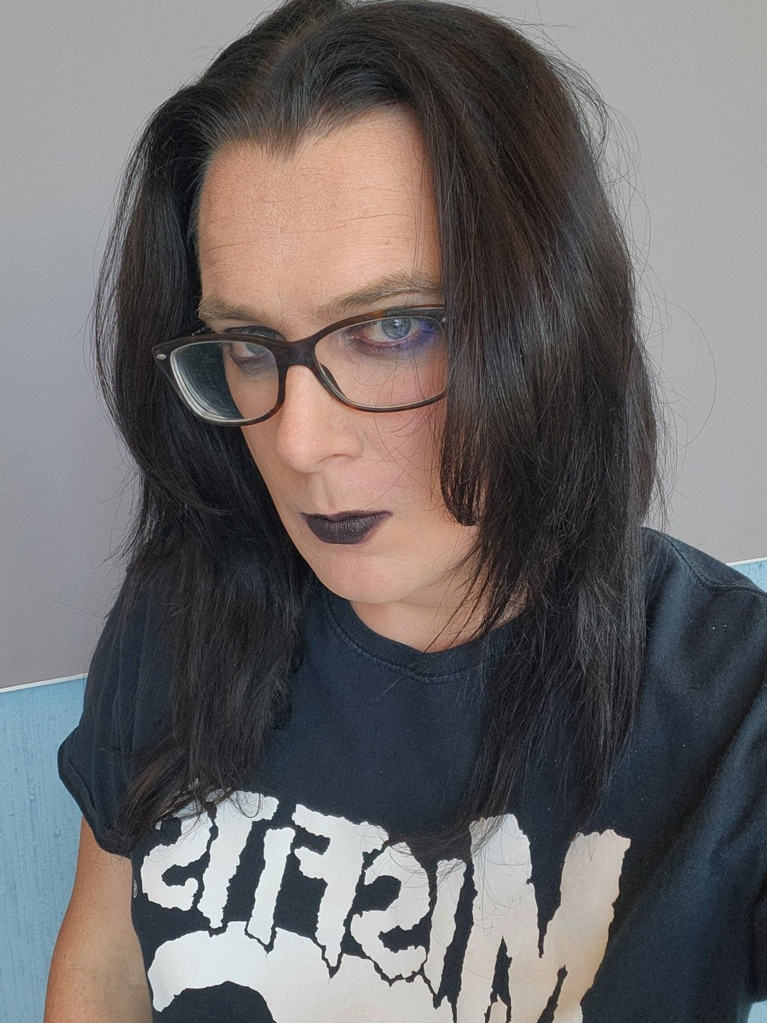 A light-skinned, non-binary person in a black misfits shirt. Their black hair is down and they have eyeglasses on. They're wearing eyeliner and black lipstick.