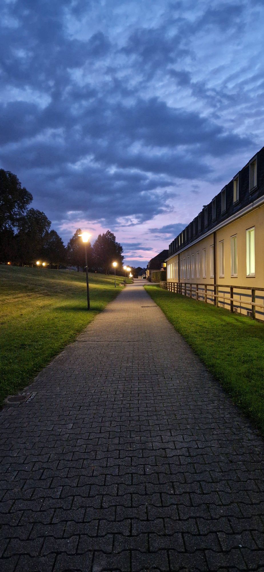 Foto. Zentral ein langer Weg, rechts ein gelber Bau, der sich über die Länge des Weges erstreckt. Es dämmert, der Himmel ist lila mit dunkelblauen Wolken. Am linken Wegesrand steht eine Straßenlaterne